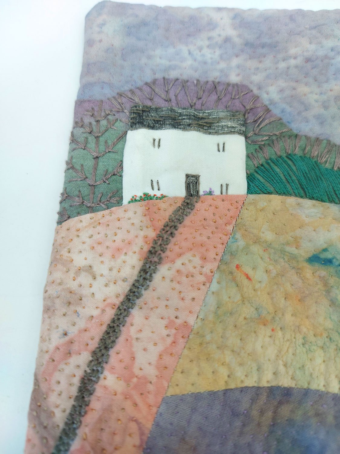 Embroidery of a white farmhouse with black roof, chimney, door, and windows, surrounded by colourful fields and trees, stitched with various textured threads.