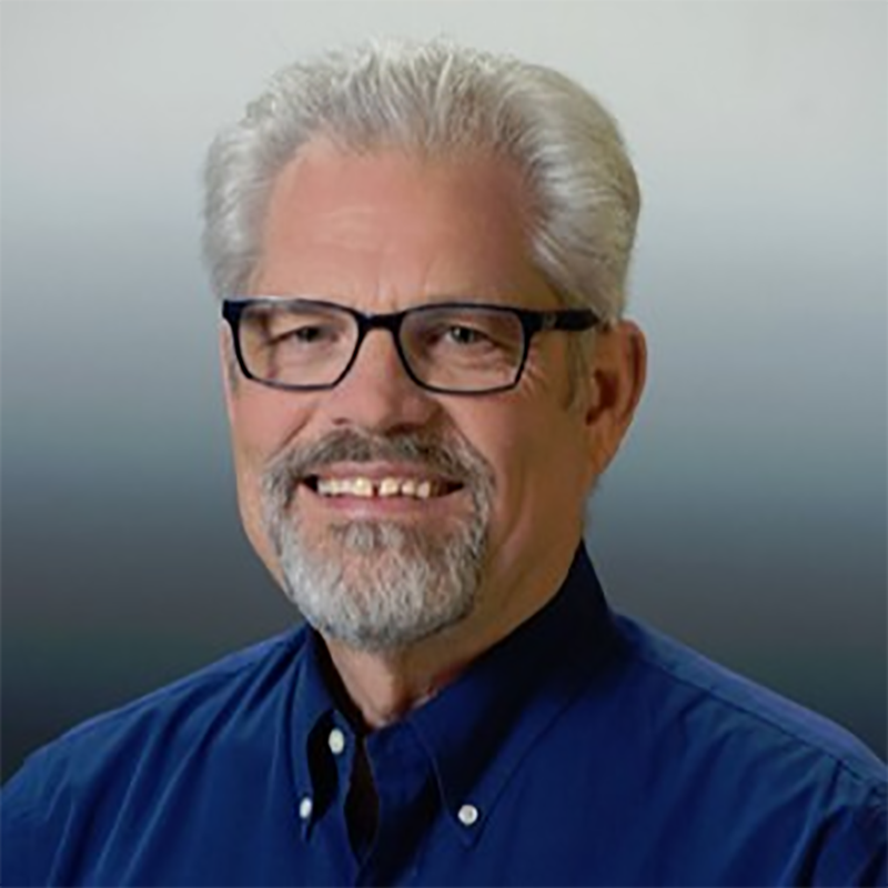 A headshot of a smiling middle-aged man with gray hair, beard, and glasses wearing a dark blue shirt against a blurred background.
