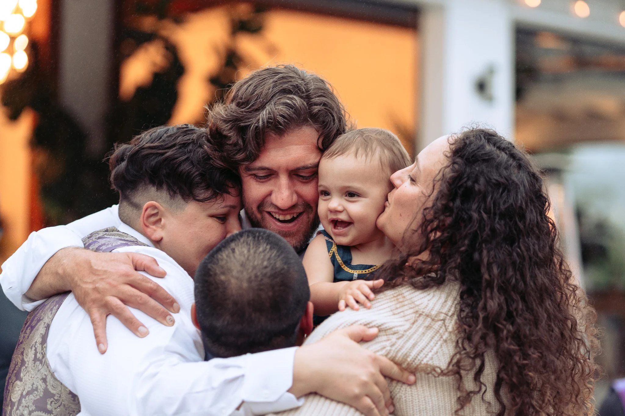 A group of people, including adults and children, are hugging and smiling warmly in an outdoor setting.