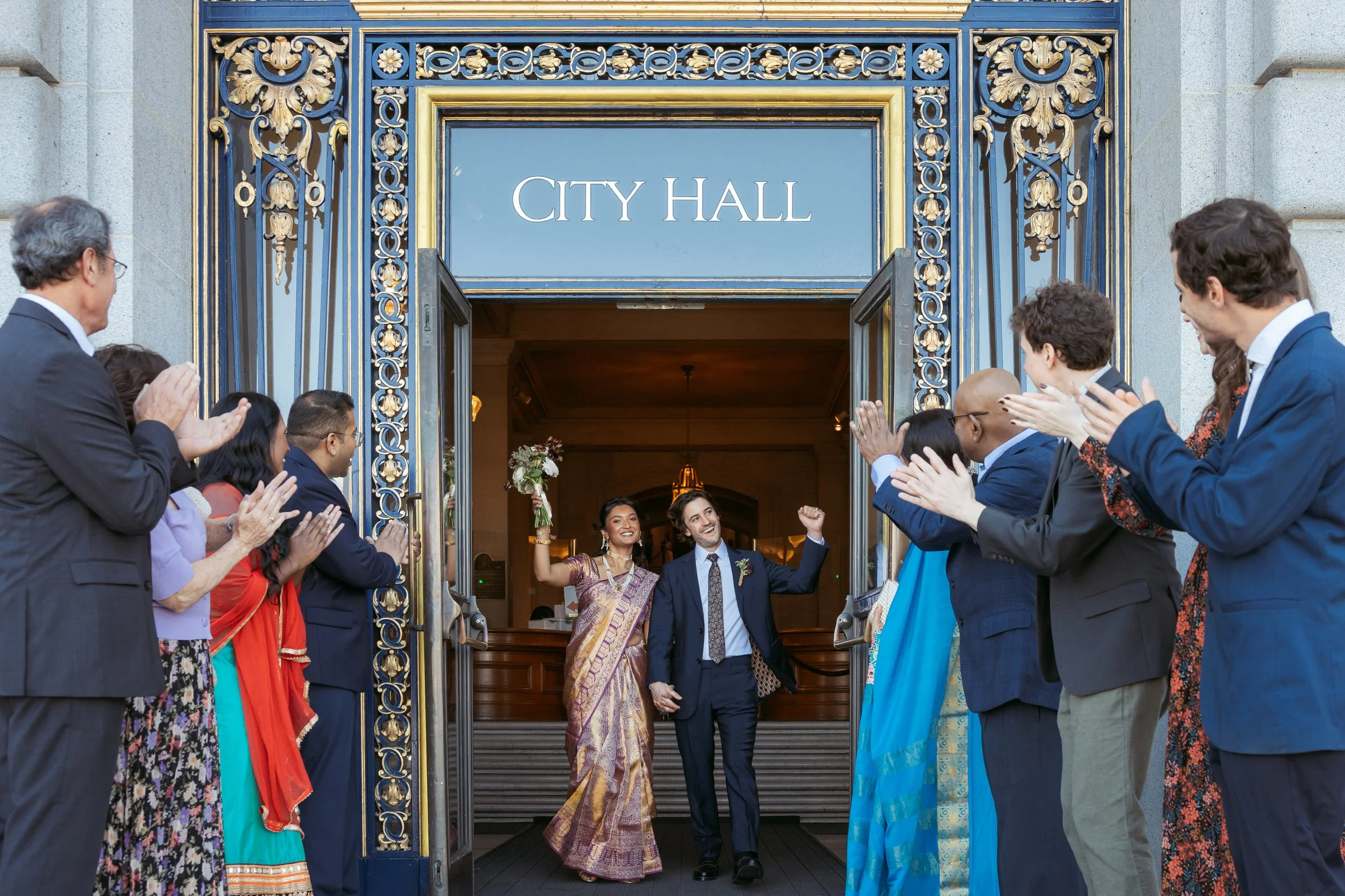 People standing outside a city hall entrance, clapping and celebrating, with a woman and a man holding hands and smiling in the center.