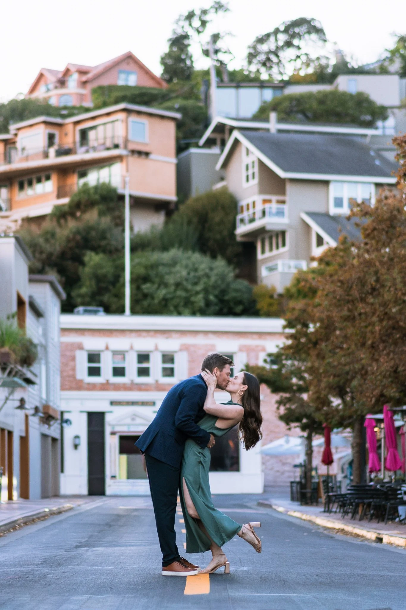 A couple kissing on the street with houses on a hill in the background