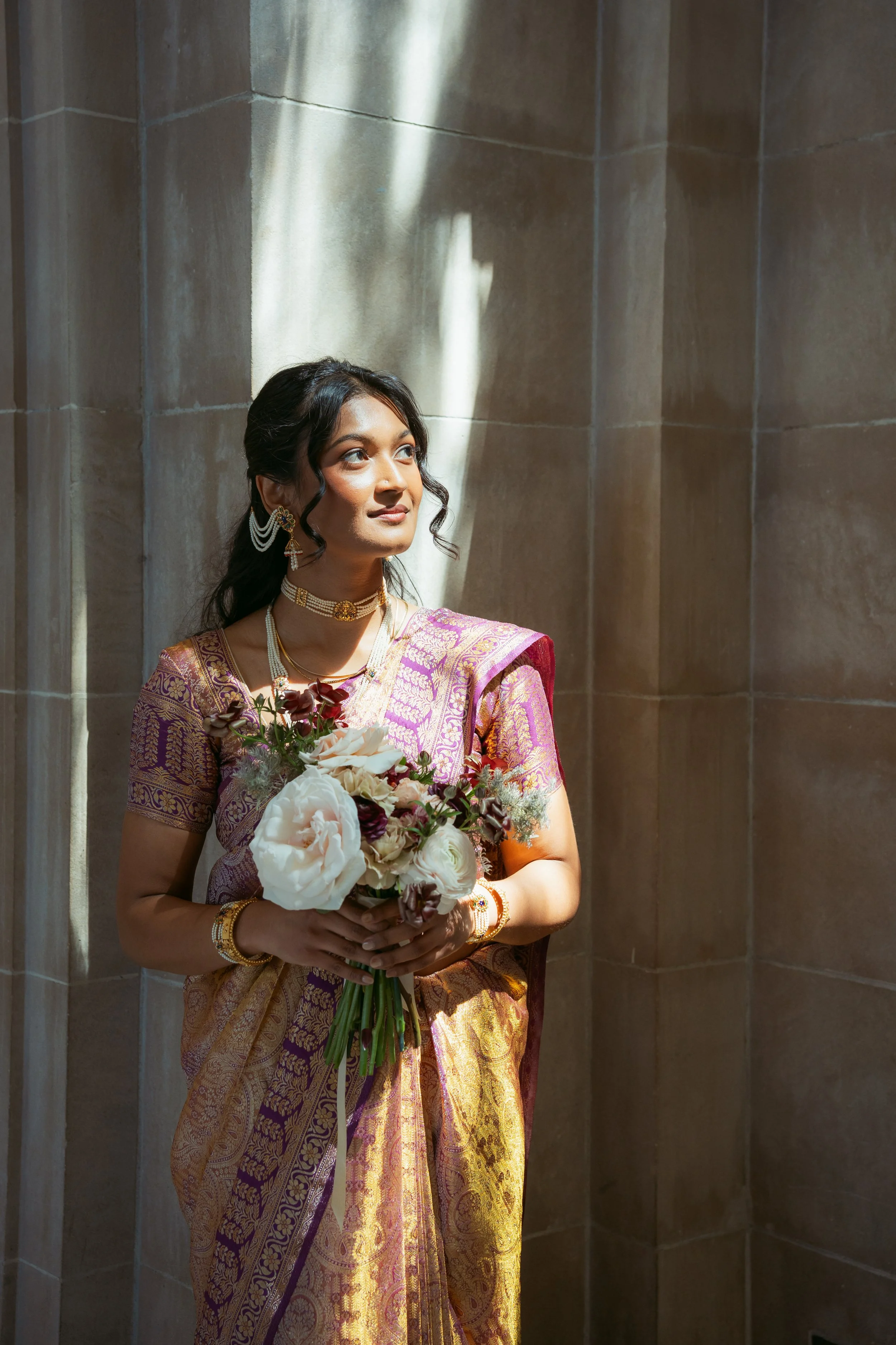 A woman in traditional Indian attire holding a bouquet of flowers, standing against a stone wall with sunlight casting a spotlight effect.