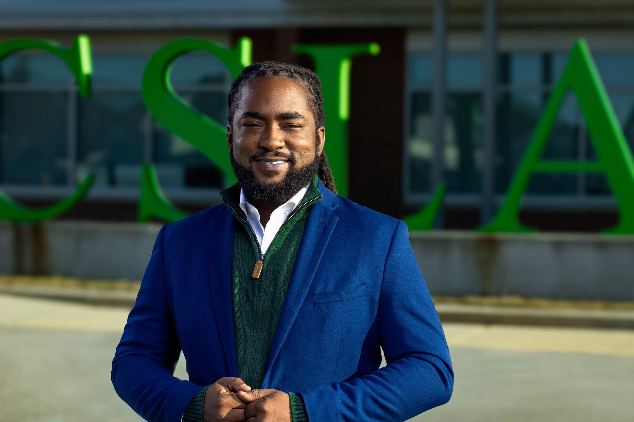 A man with braided hair and a beard standing outside in front of a building with large green letters.