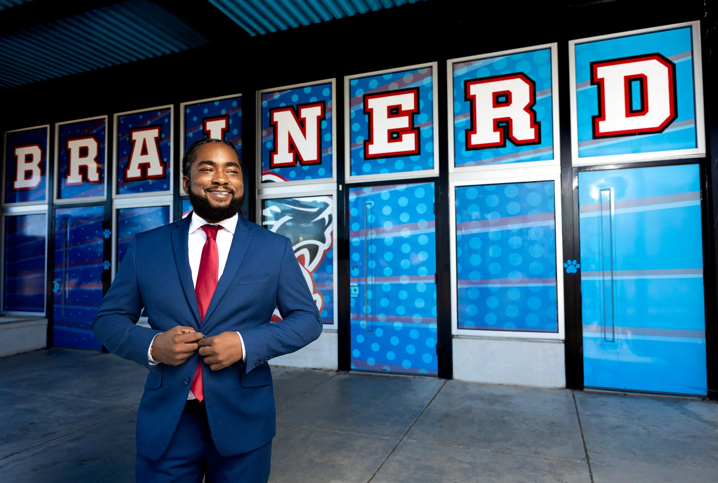 A man in a navy blue suit with a red tie standing outside a building with a sign that spells 'BRANDNER' in large red and white letters with a blue background.