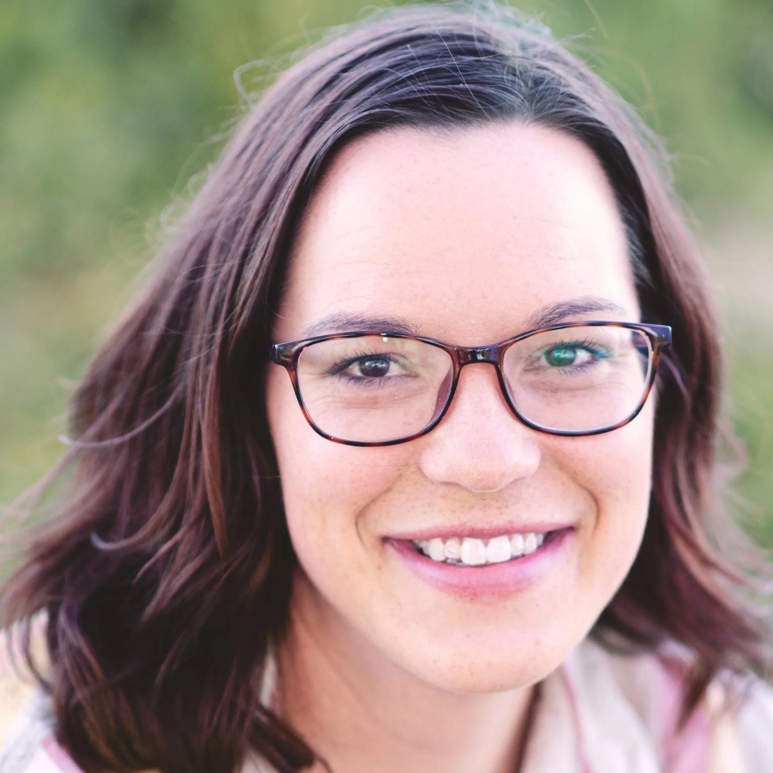 Close-up image of a woman smiling, wearing glasses, with brown hair, outdoors with blurred green background.