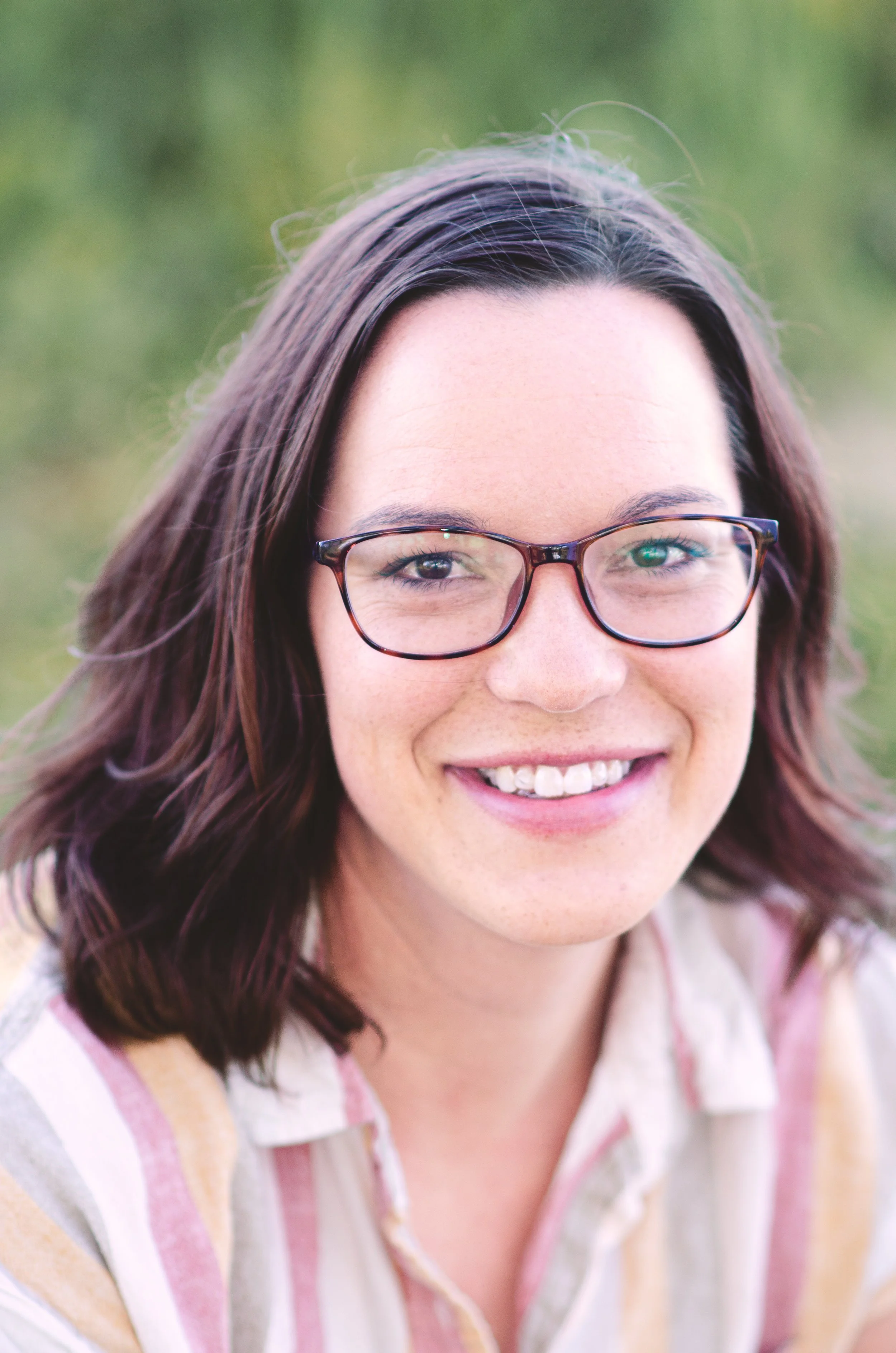 Close-up of a woman with shoulder-length wavy brown hair, wearing glasses, smiling, outdoors with blurred green background.
