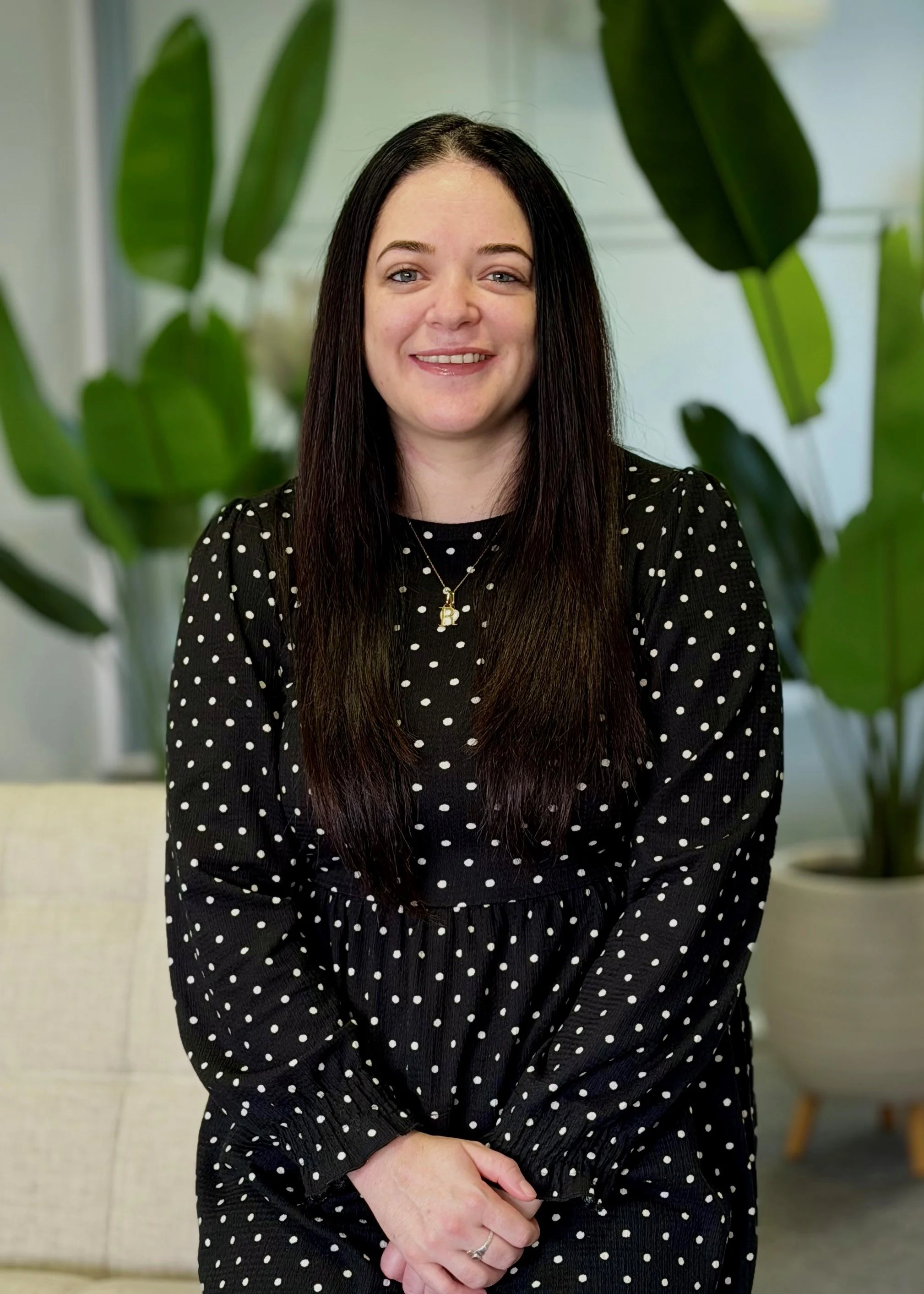 A woman with long black hair, wearing a white blouse with ruffled cuffs and collar, and a black pinafore dress, sitting inside near large green plants and smiling at the camera.