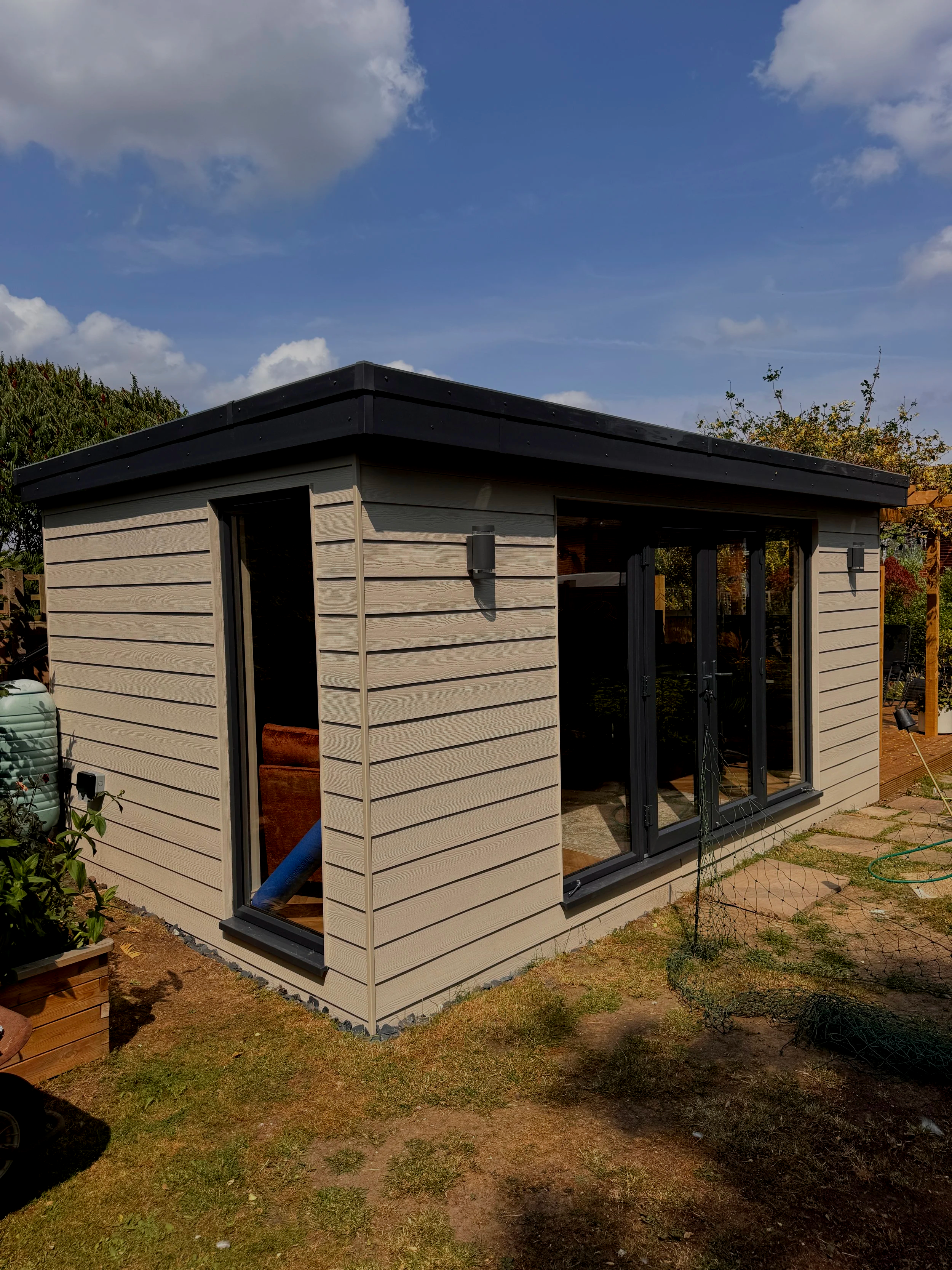 A modern garden room with black-framed glass doors and beige horizontal siding outside on a partly cloudy day.