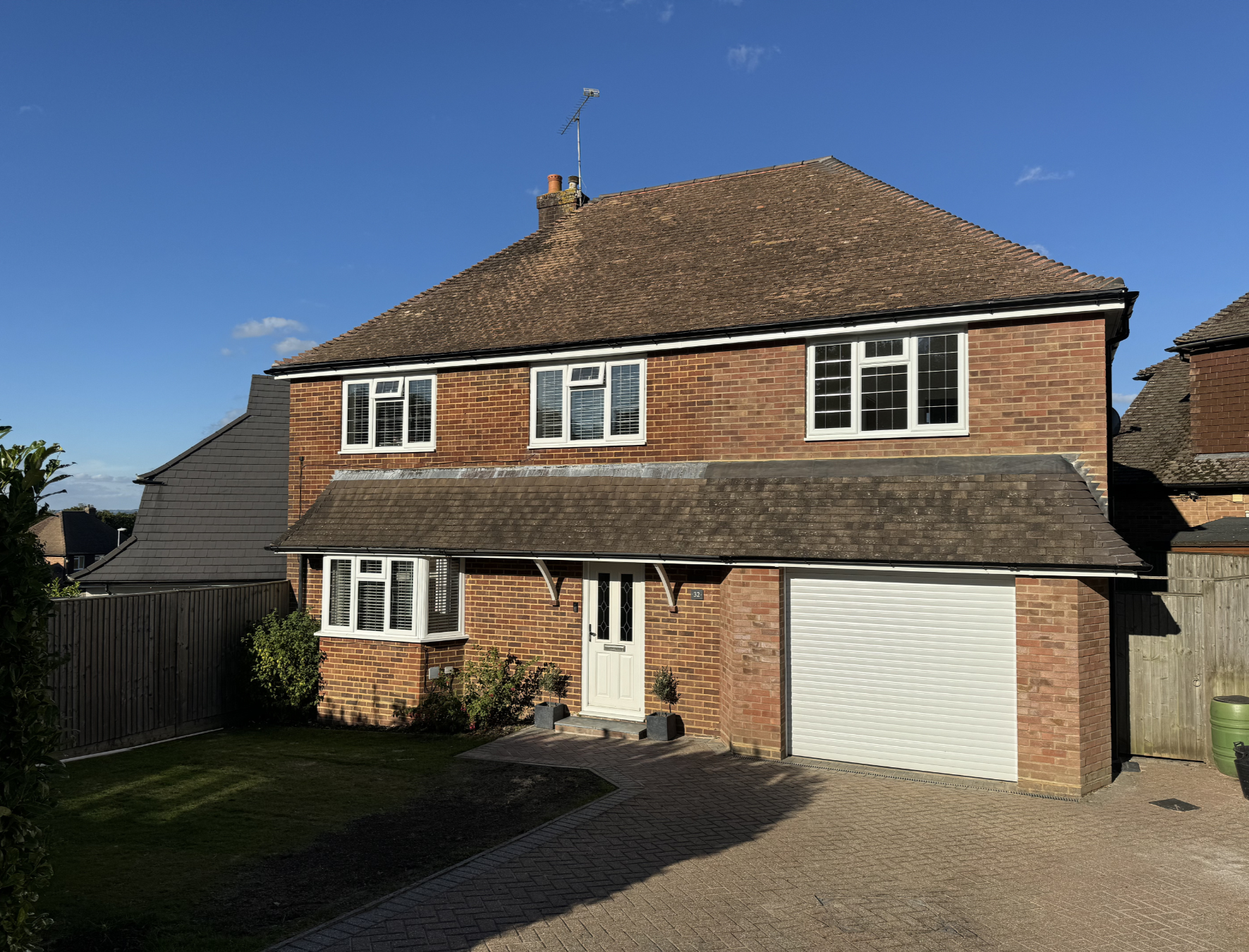 A two-story brick house with a pitched roof, white-framed windows, and a white garage door, with a small garden and paved driveway in front under a clear blue sky.