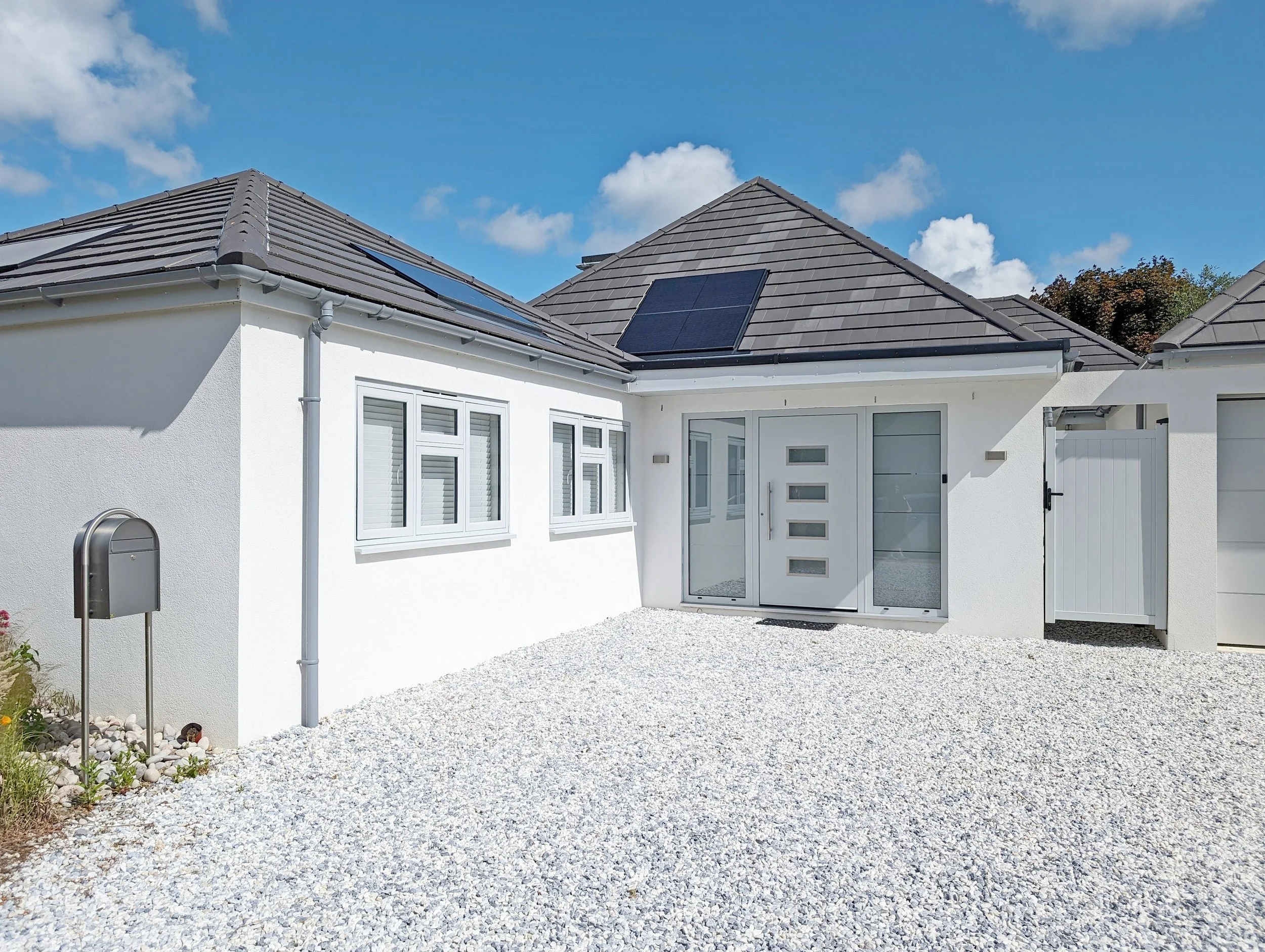 A modern white house with a gray tiled roof, solar panels, and a gravel yard under a blue sky with clouds.