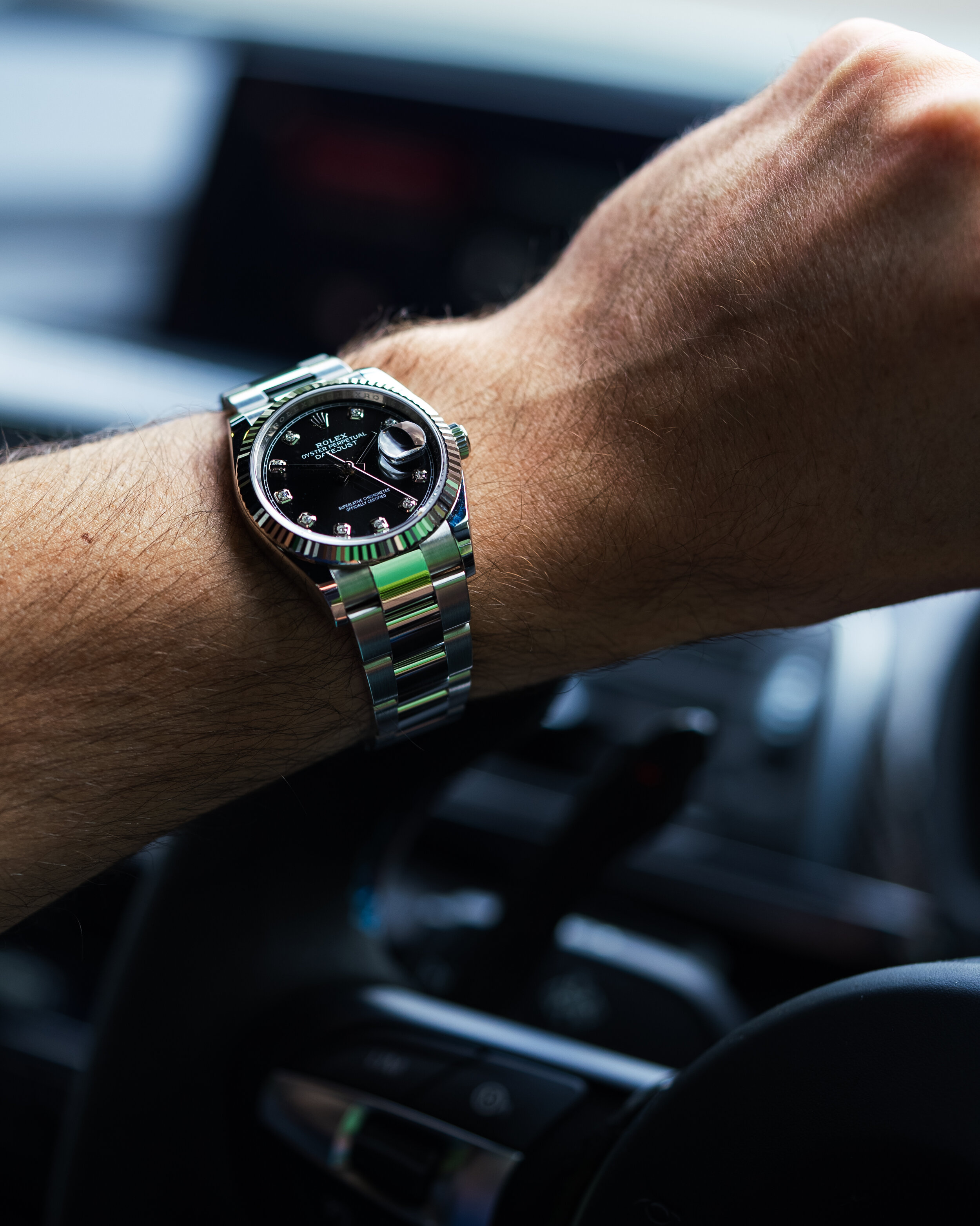 Wrist wearing a Rolex watch with a black dial, silver and diamond markers, inside a car.