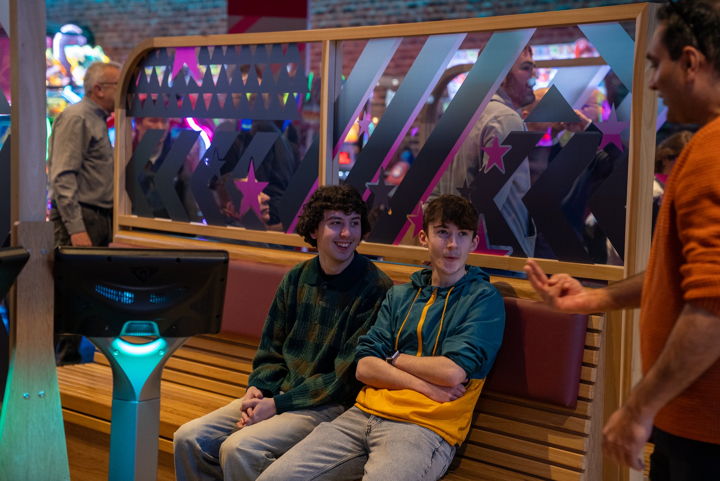 Two young people sitting on a bench in an indoor gaming arcade, with a decorative divider and arcade machines in the background.
