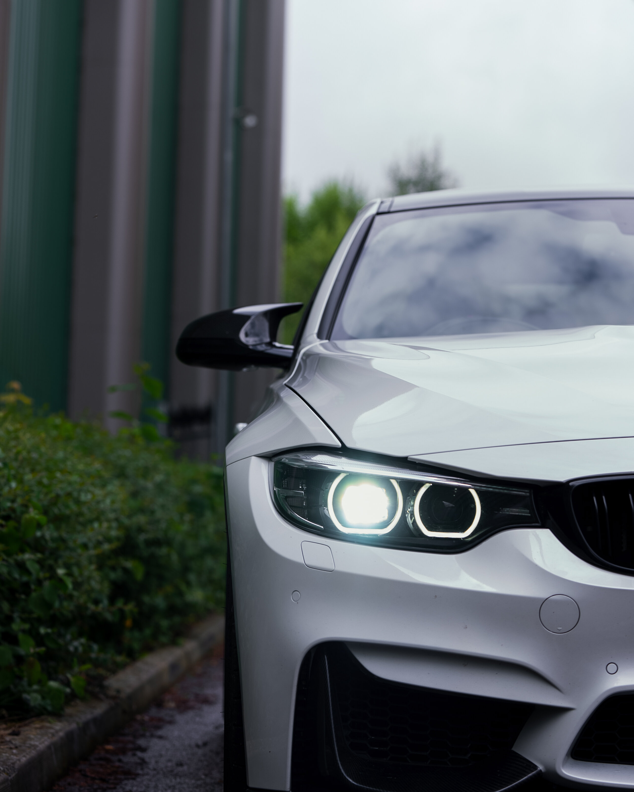Close-up of a white car's front with illuminated headlights, side mirror, and part of the grille visible, set against greenery and a building background.