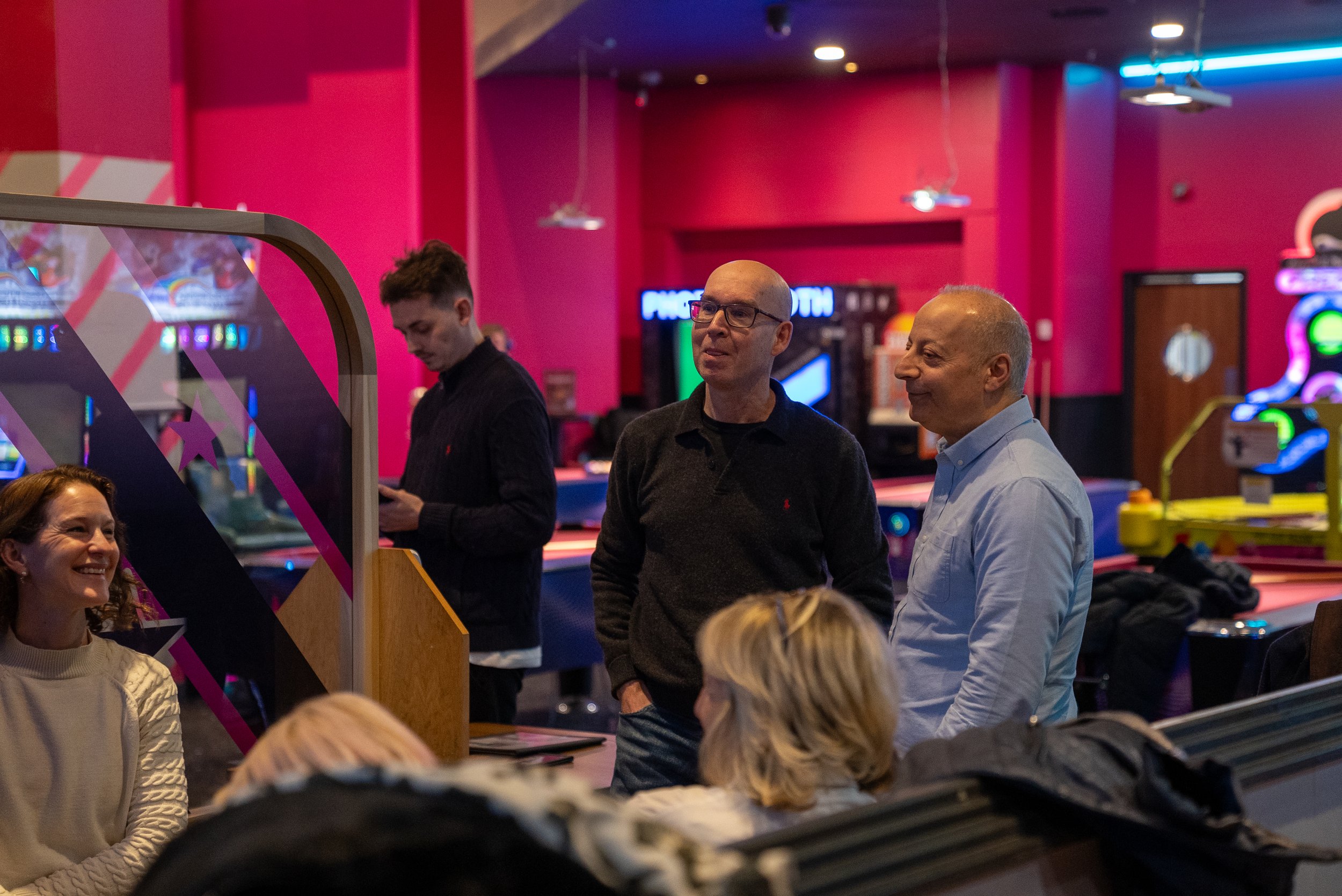 People socializing in an indoor entertainment venue with arcade games and pink lighting.