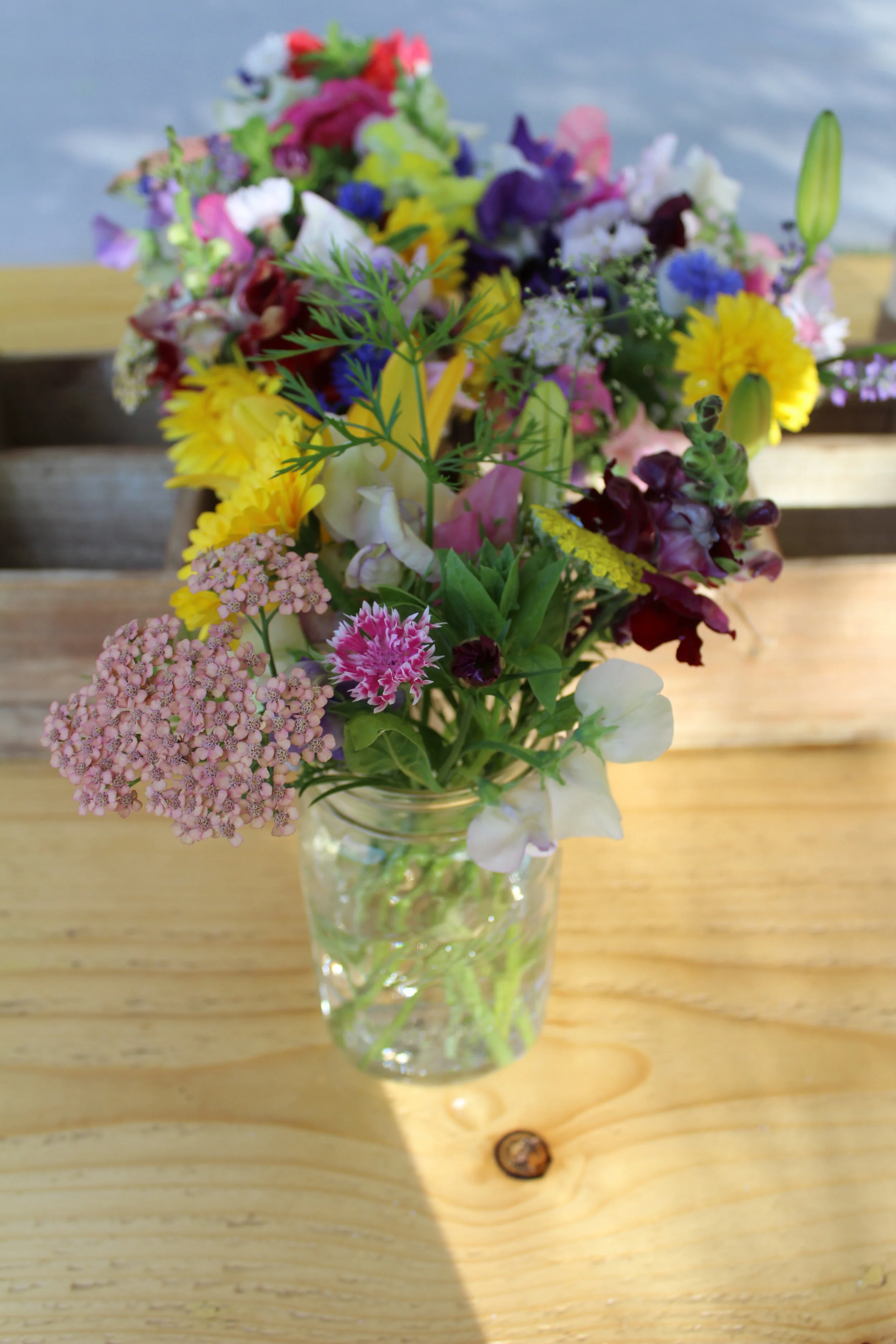 A colorful bouquet of various flowers in a clear glass jar on a wooden table.