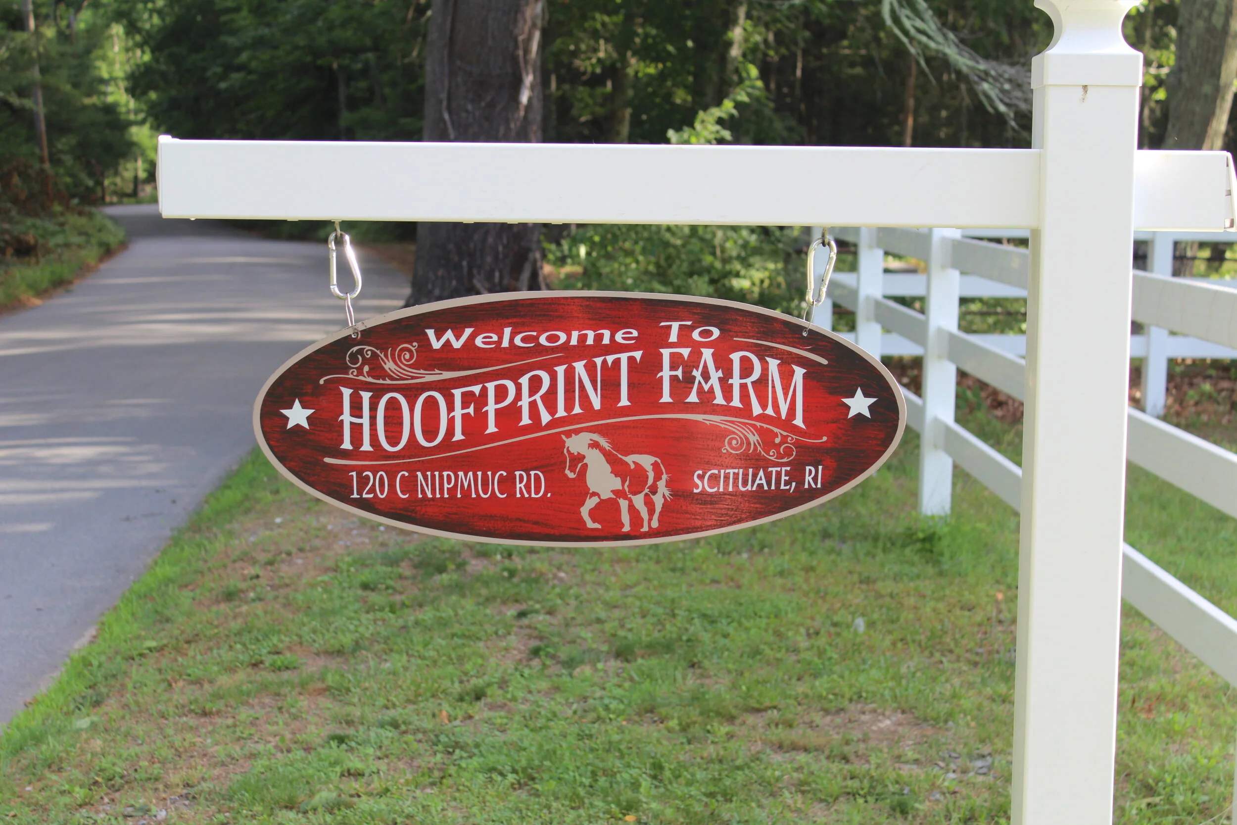 Sign for Hoofprint Farm, 120 C Nipmuc Rd, Scituate, RI, with a decorative horse graphic, hanging on a white post next to a road.