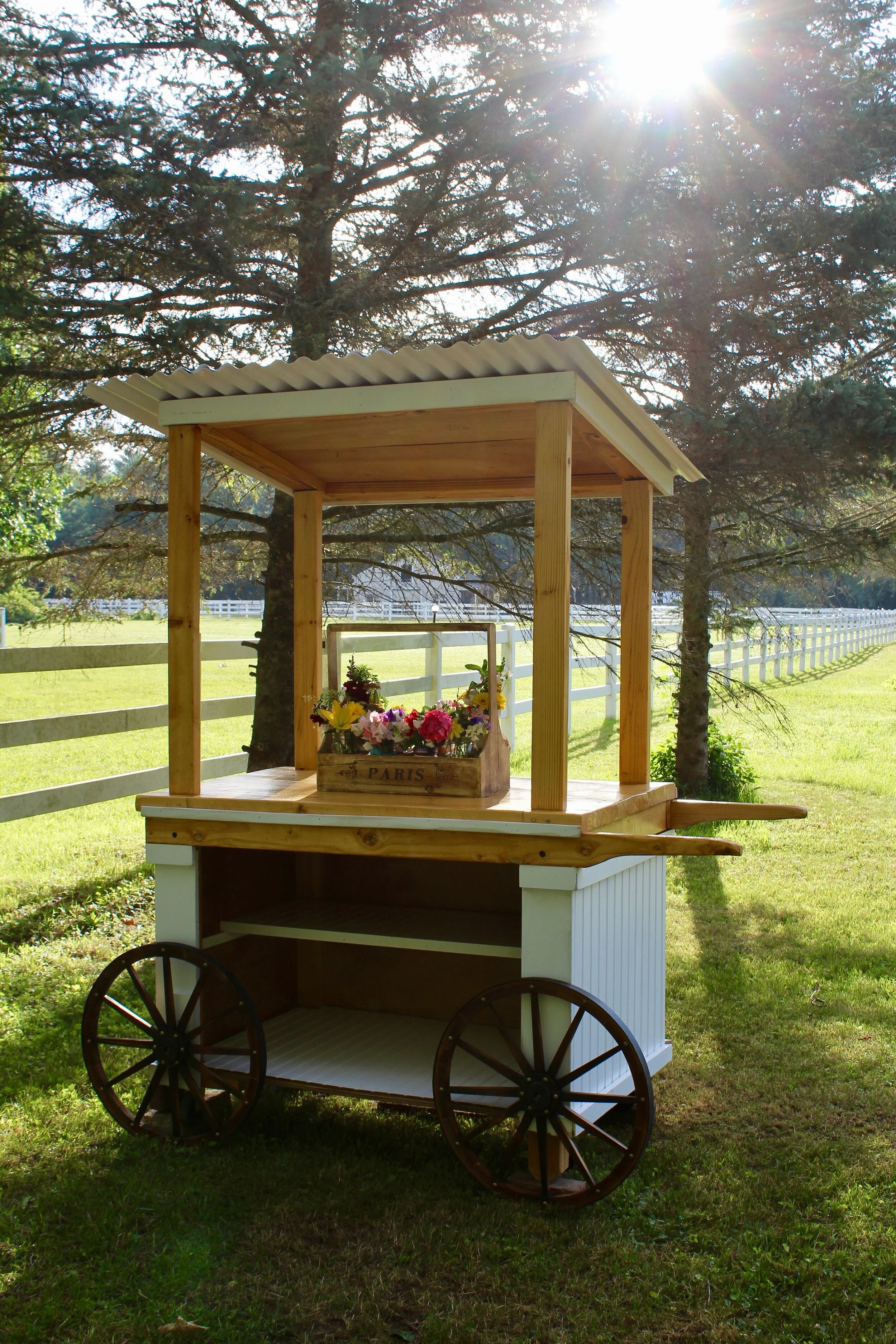 Wooden flower cart with wheels and a canopy, holding a crate of colorful flowers, set in a grassy outdoor area with trees and sunlight streaming through.