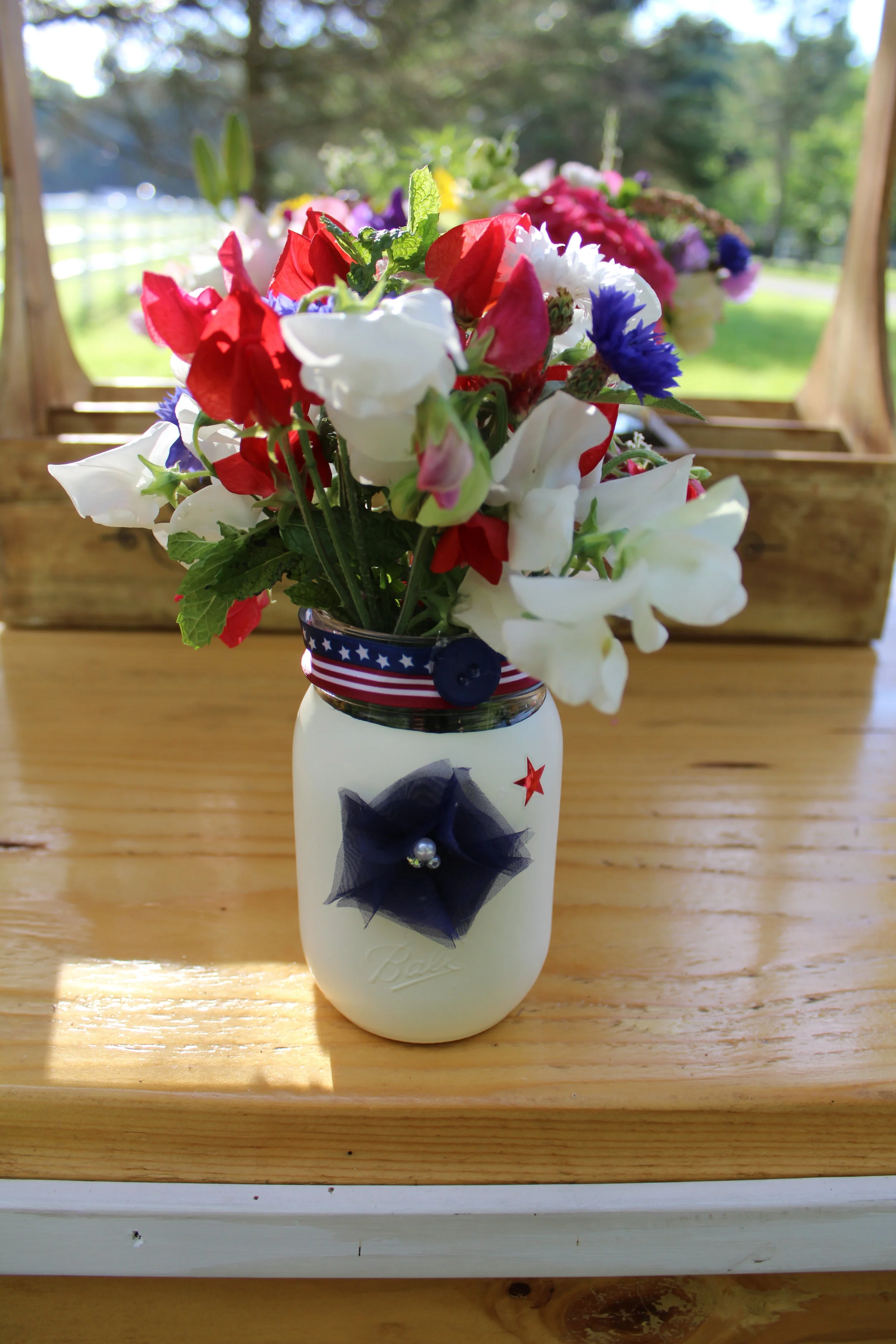 A white mason jar decorated with a navy blue ribbon and star, filled with red, white, and blue flowers, standing on a wooden table with a garden in the background.