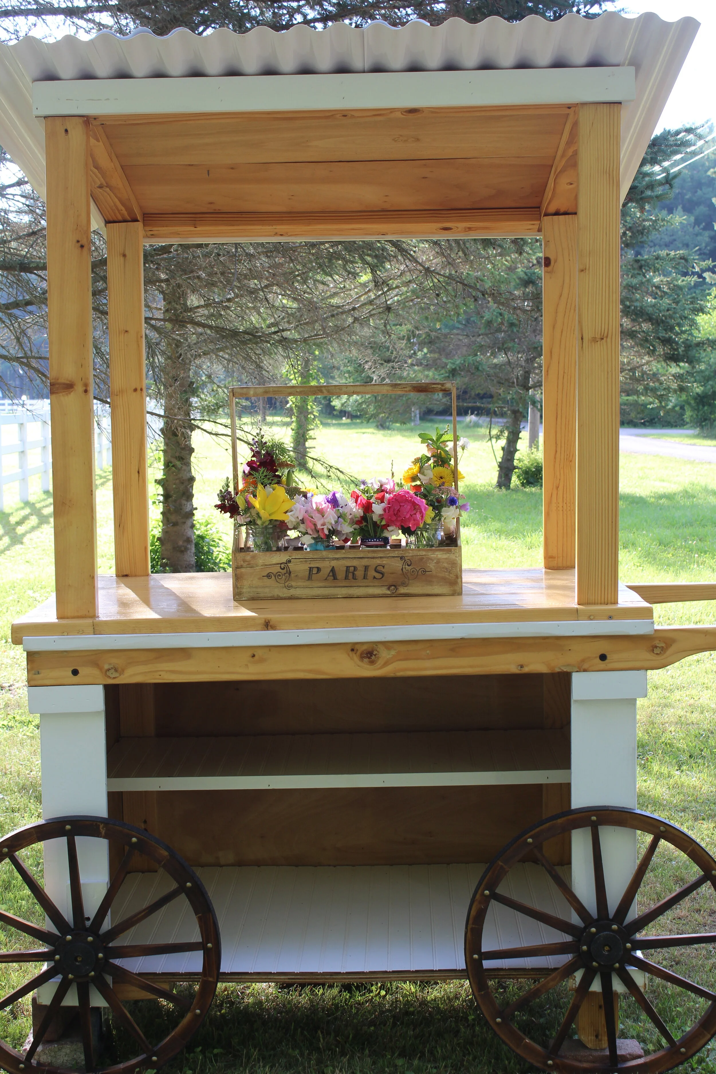 Wooden cart with corrugated roof, featuring flowers in a "Paris" box, surrounded by trees and grassed area.