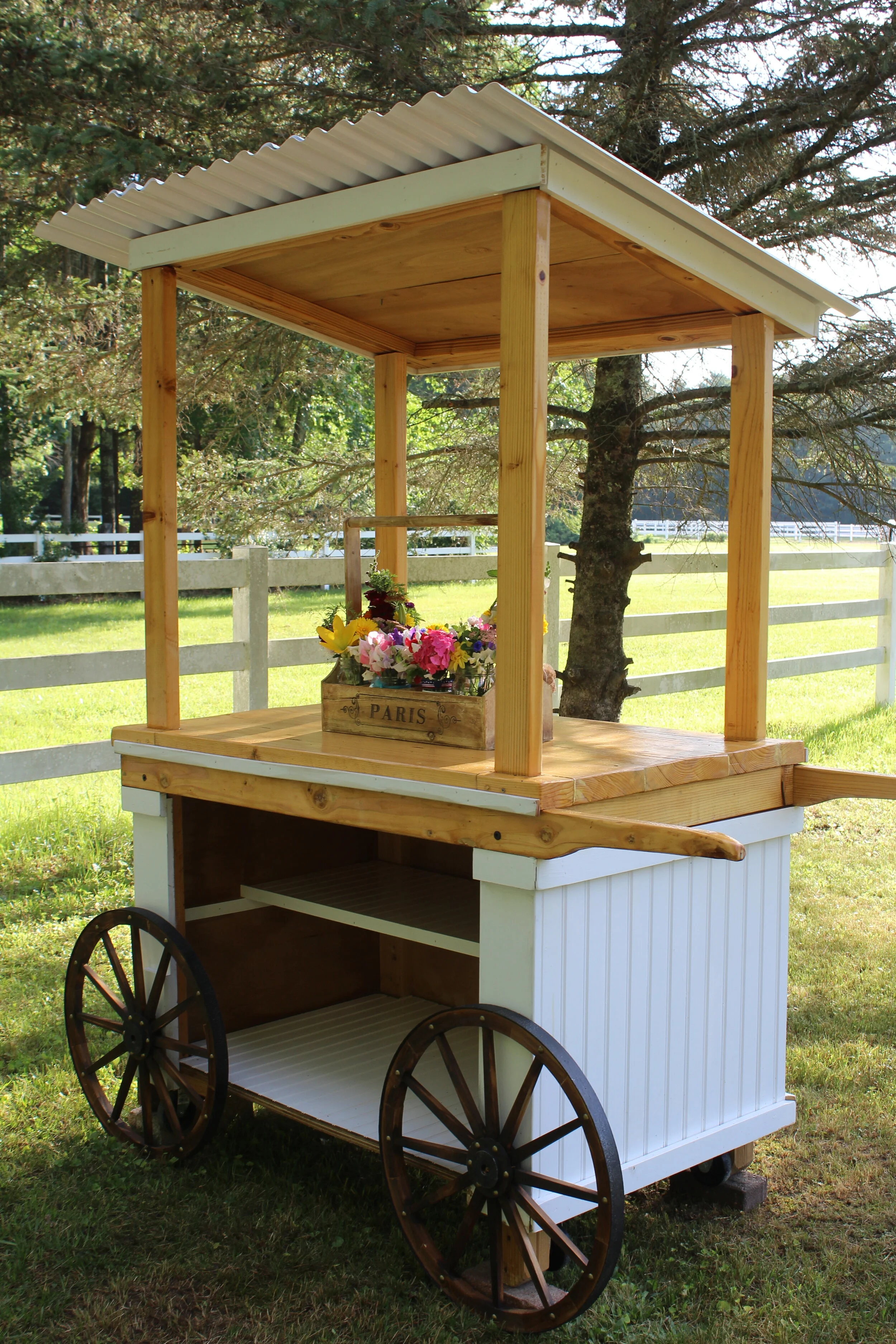 Wooden flower cart with wheels, a corrugated roof, and a crate of colorful flowers. Set outdoors in a grassy area with trees and a white fence in the background.
