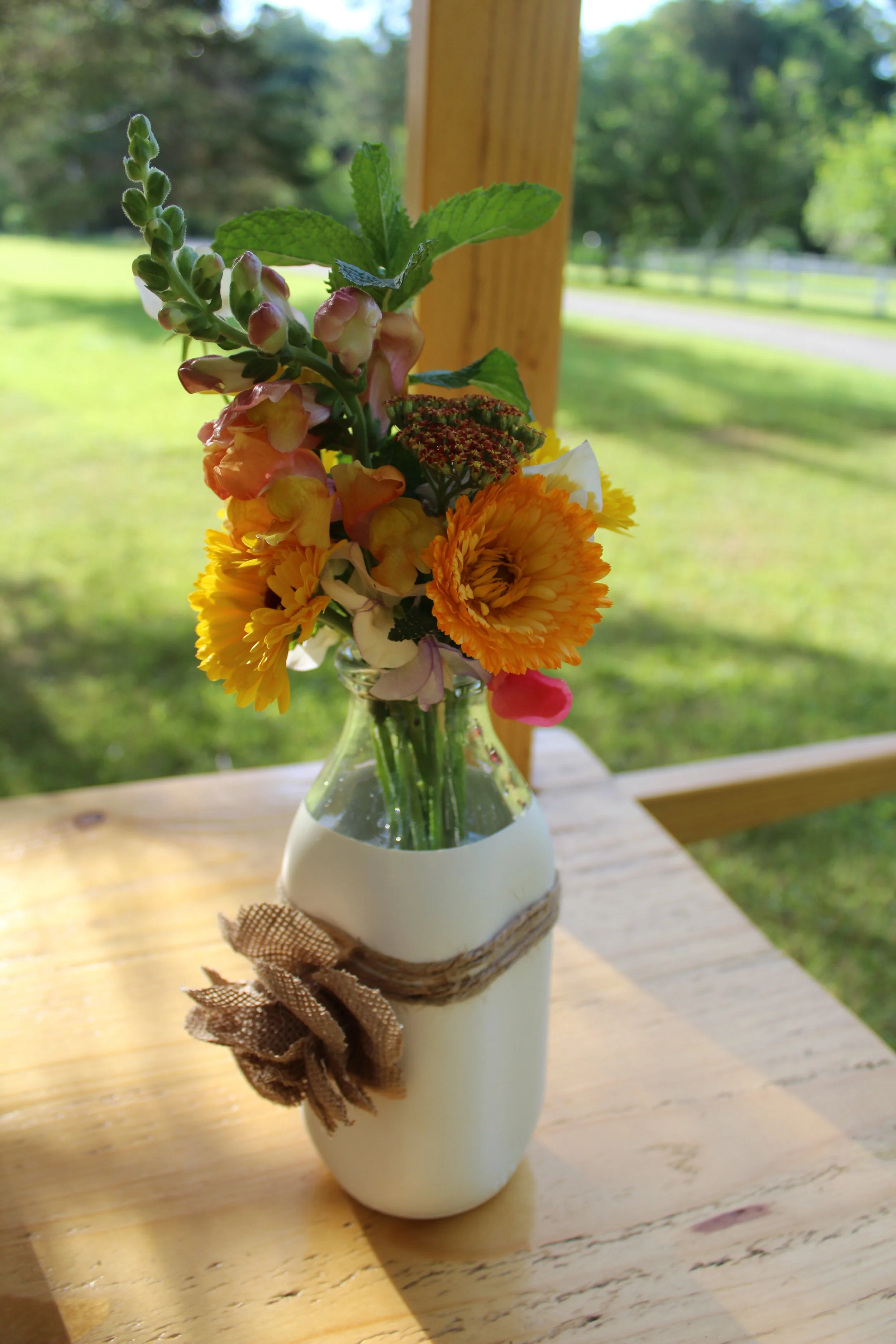 A bouquet of colorful flowers in a decorative white vase with a burlap ribbon on a wooden table outside.