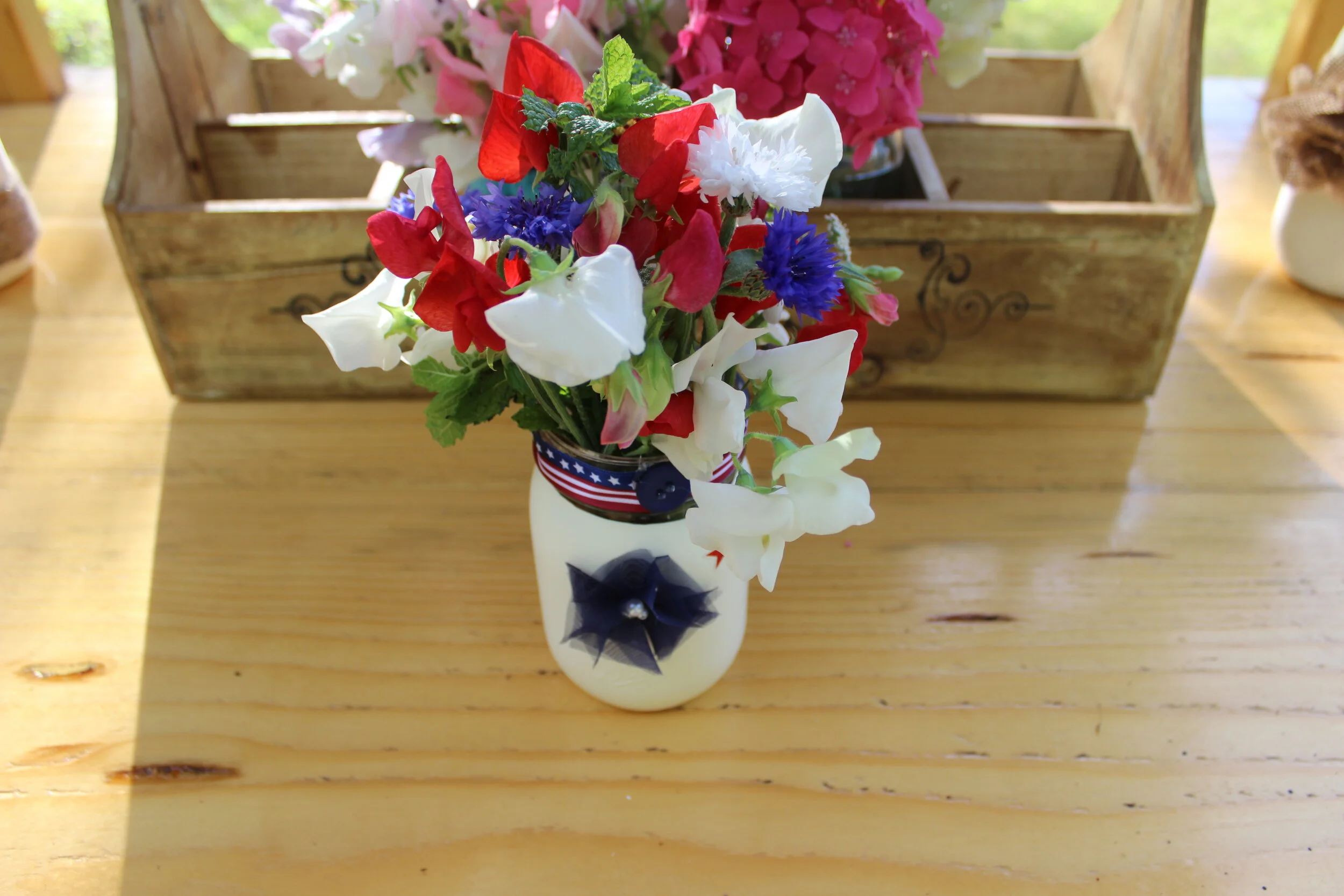 Patriotic-themed flower arrangement in a white jar with red, white, and blue flowers, decorated with ribbon on a wooden table.