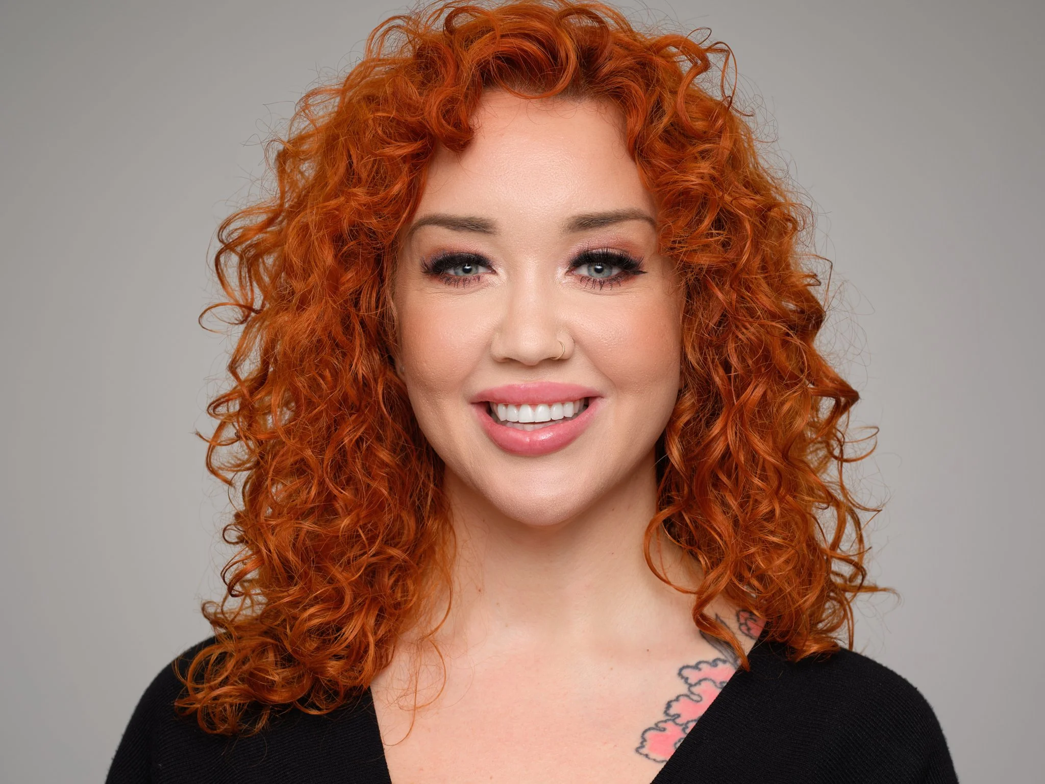 Portrait of a woman with curly red hair, blue eyes, sitting against a neutral gray background, wearing a black top and a tattoo on her chest.