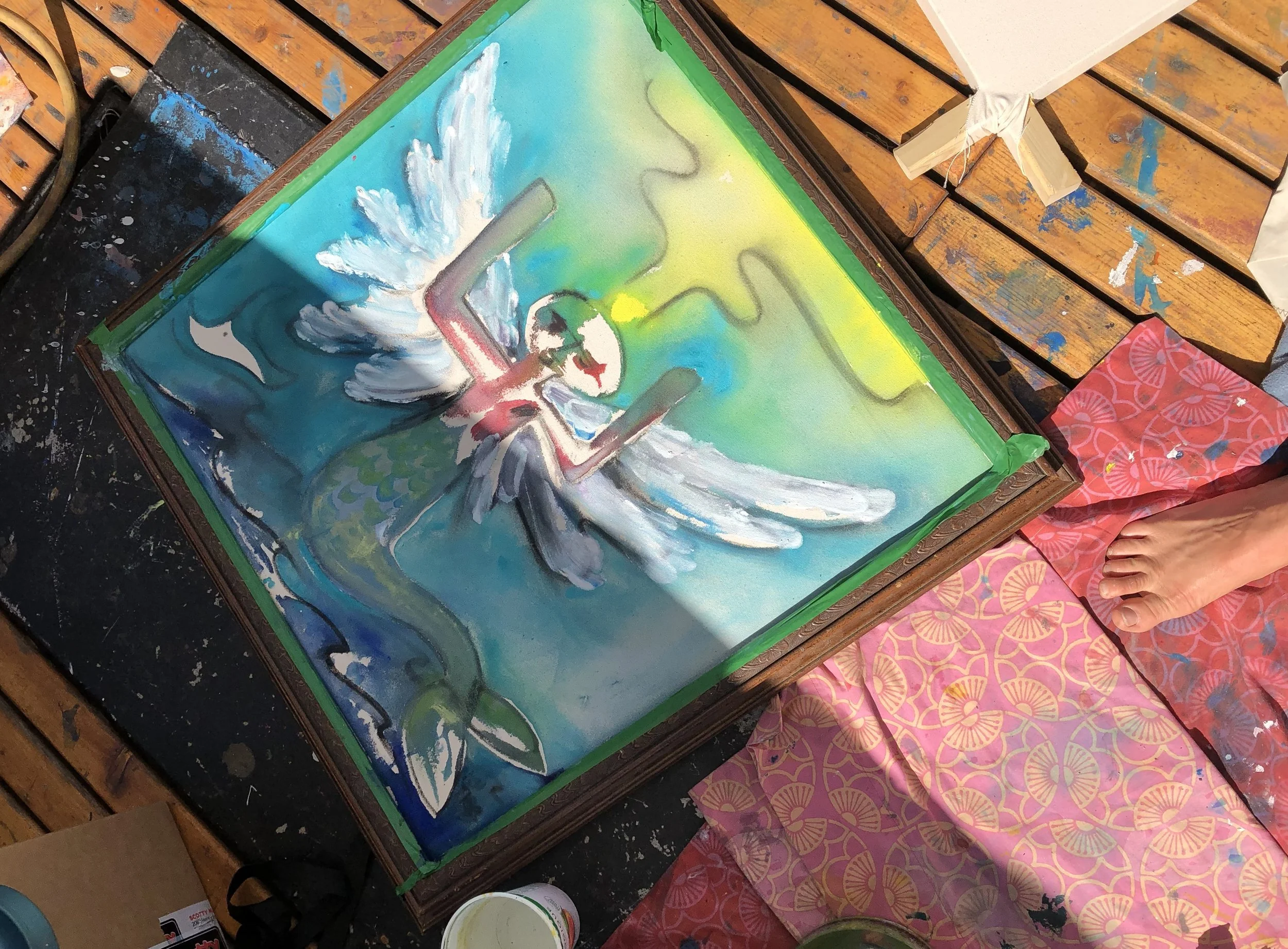 Close-up of a painting on a wooden table with green tape around the edges. The artwork features a dove with outstretched wings, a pipe, and an abstract background with green, yellow, and blue tones. Surrounding the table are painting supplies, includ