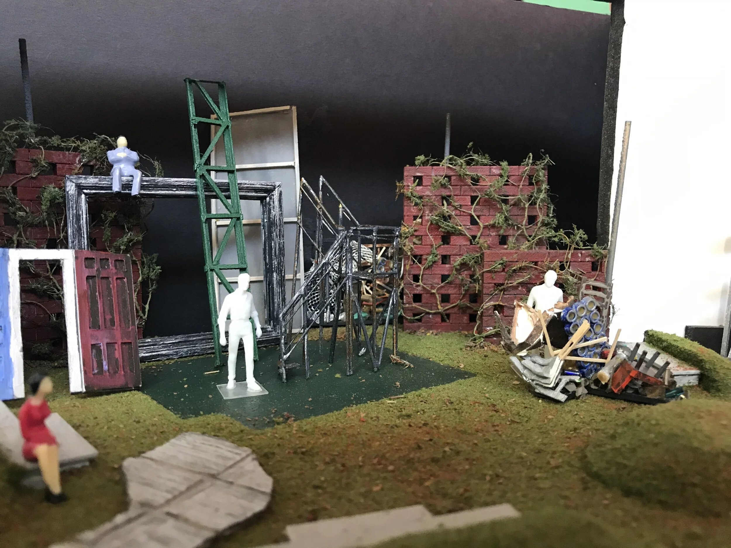Model by: Melpomene KatakalosAn Astroturf floor supports brick walls covered in vines and other architectural flotsam. At center, a truss leans like a ladder against a large picture frame. A plastic figure, painted blue, sits on top. At left sits a plastic figure, dress painted red, and shoes painted black. At center stands an unpainted, plastic figure, all white. At right stands another unpainted, plastic figure, all white, visible from the waist up behind a pile of junk.