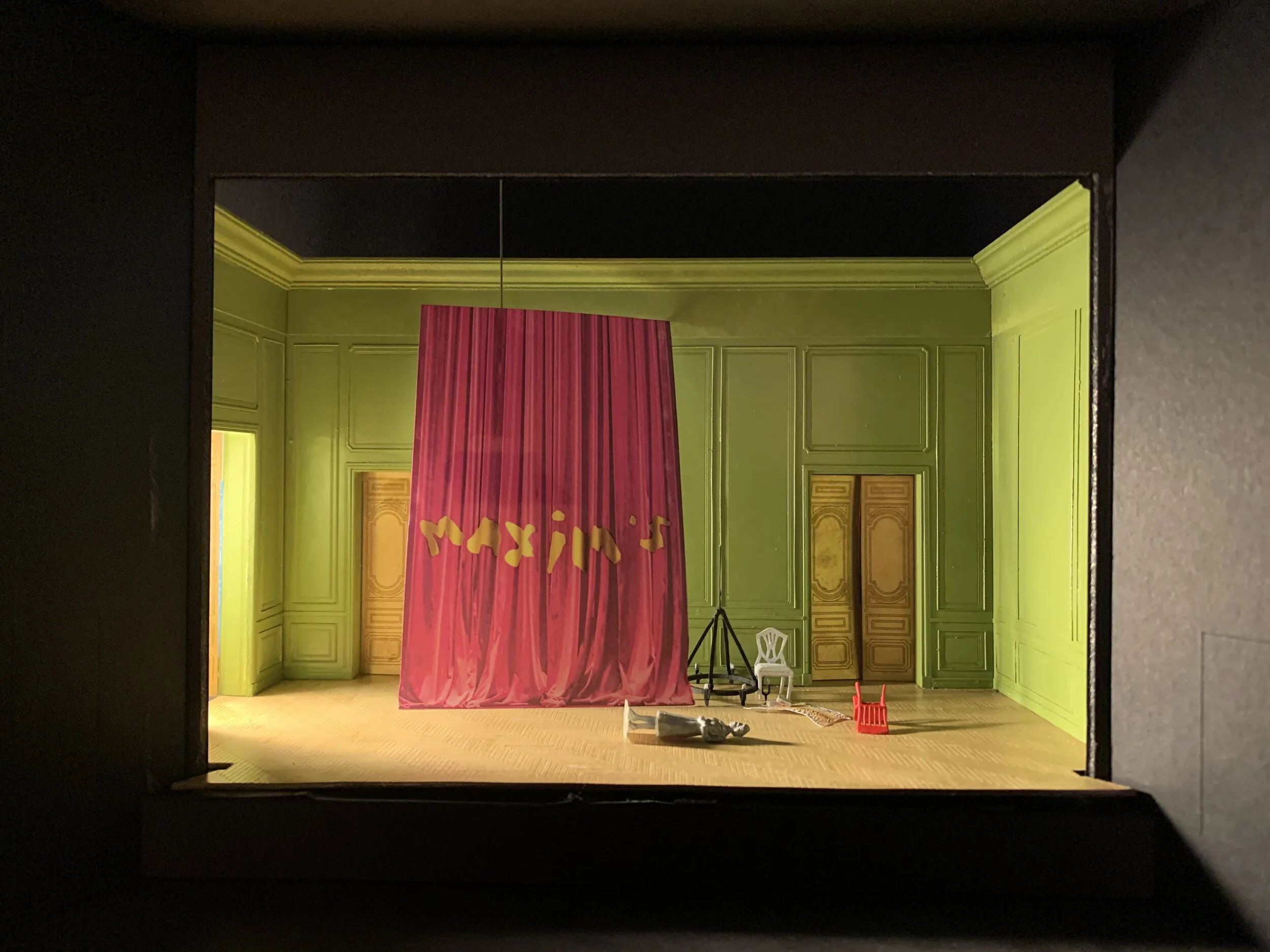 Model by: Alexander WoodwardThree, tall, lime-green paneled walls with yellow doors describe a room. Suspended in the middle is a gathered curtain of magenta fabric that reads “Maxim’s”. Some remnants including a white chair, an upended red chair, and a gray, plastic figure supine on the floor.