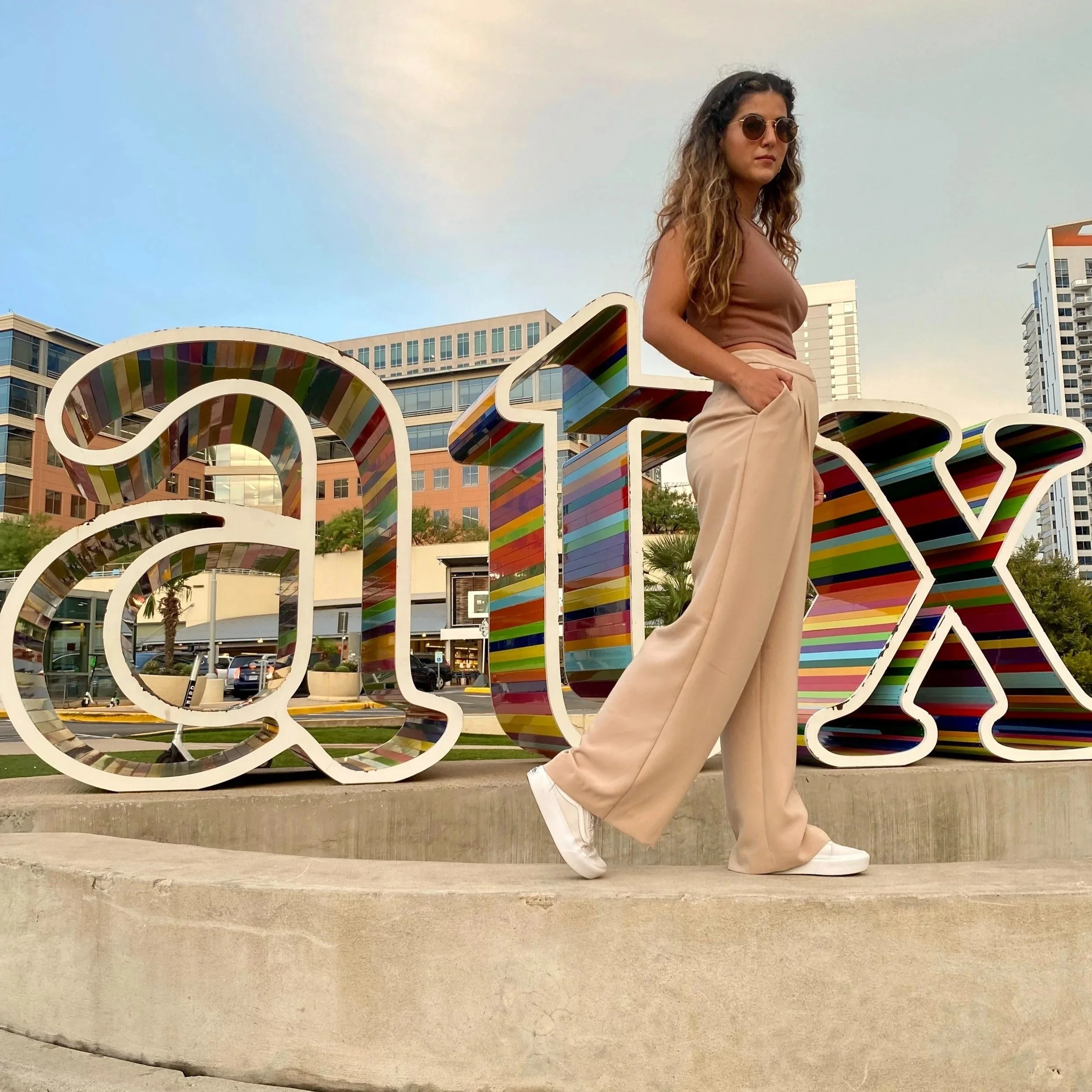 Woman standing next to colorful 'art' sculpture in city park in Austin, wearing beige pants, brown top, white sneakers, sunglasses, with tall modern buildings in background during daytime.