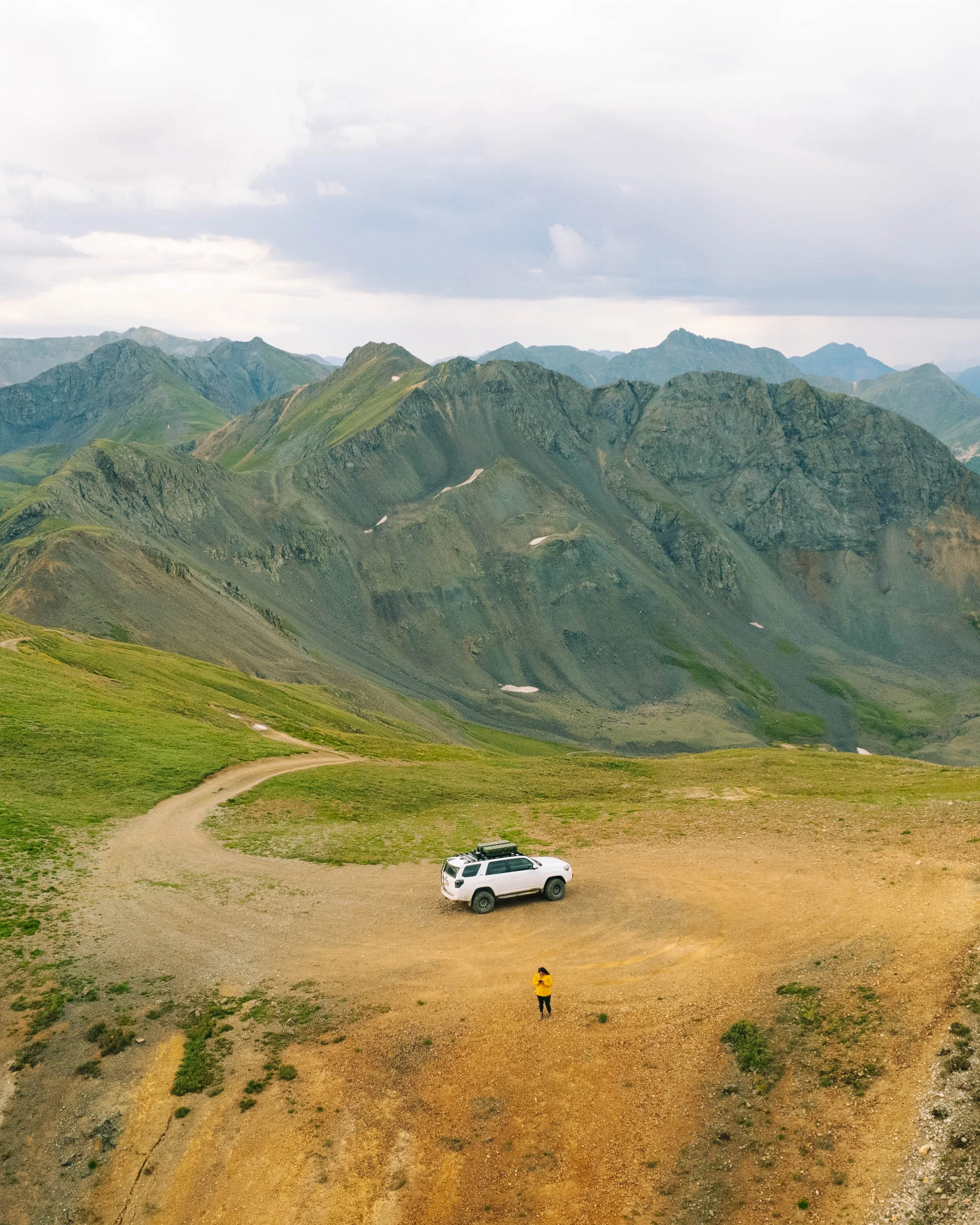 A person in a yellow jacket stands on a dirt trail next to a white SUV in a mountainous landscape with green hills and rugged mountain peaks in the background.