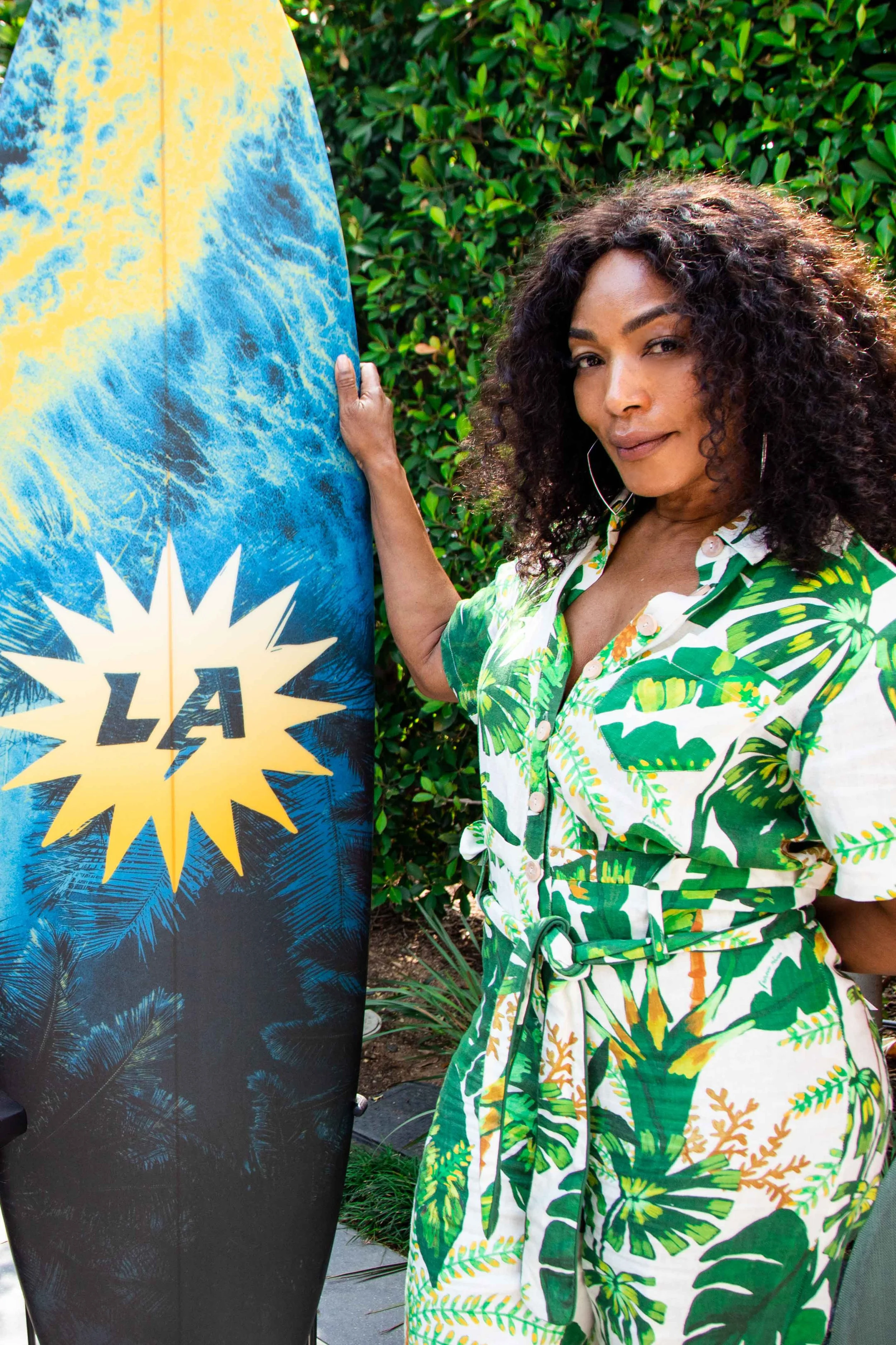 A woman with dark curly hair wearing a green and white tropical print dress standing outdoors next to a surfboard with a blue, yellow, and black design and the word 'LA' on it, with green foliage in the background.