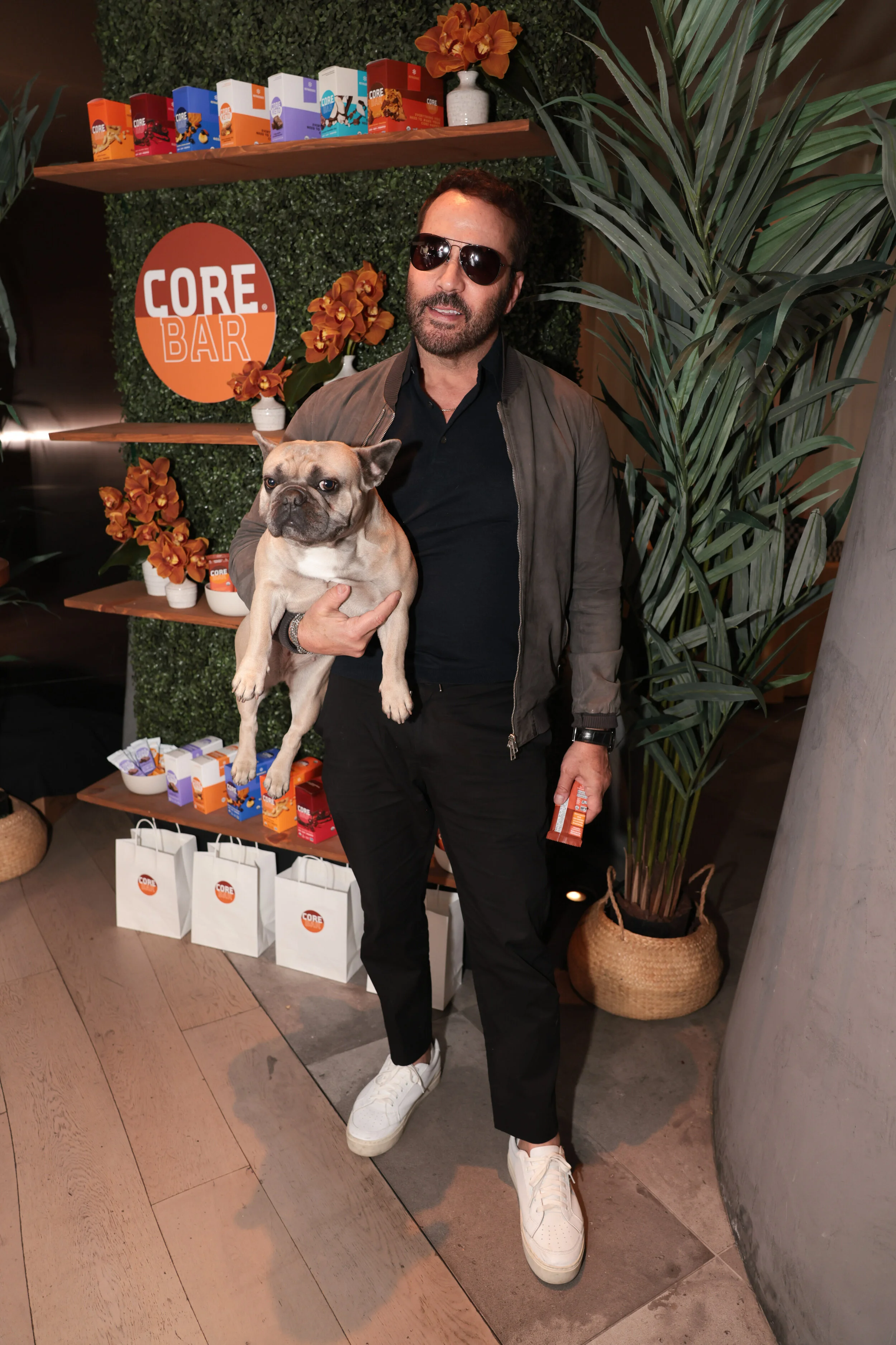 A man wearing sunglasses holding a French Bulldog in front of a display with a sign that reads 'Core Bar', decorated with orange flowers and green foliage, at an indoor event.
