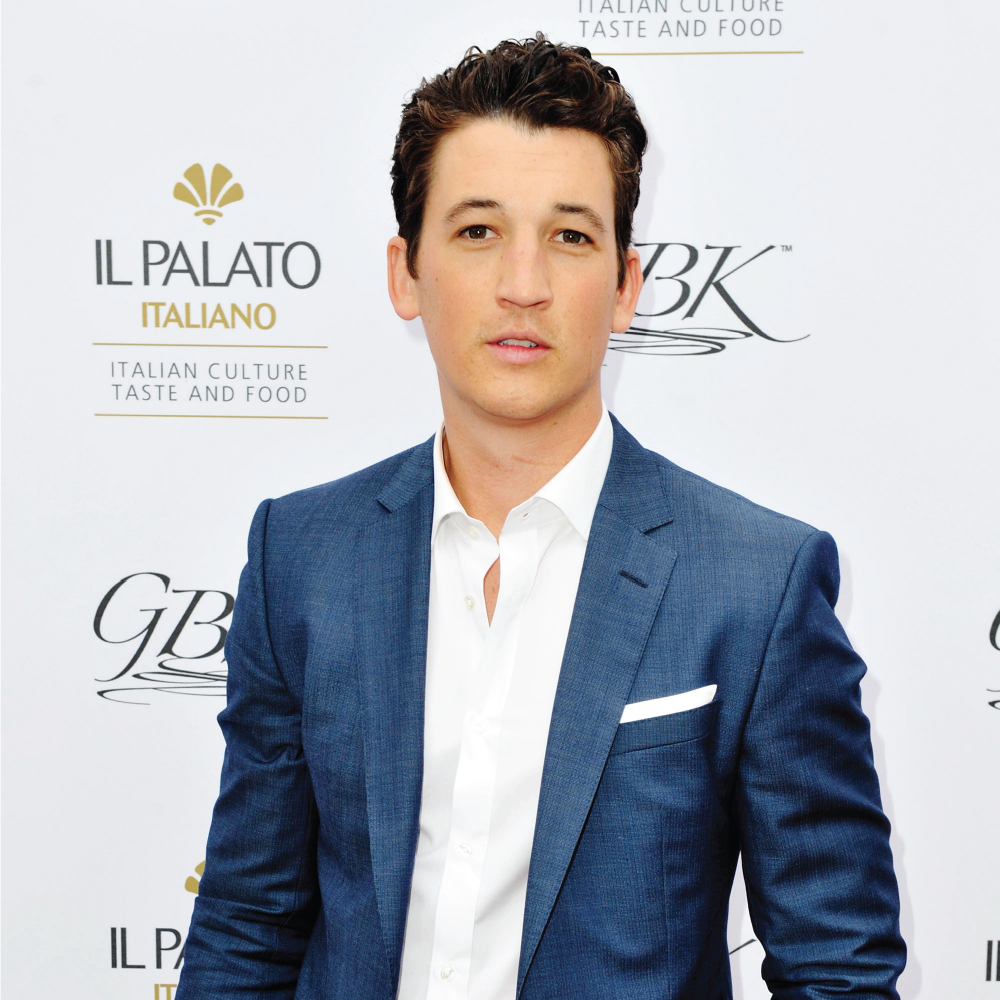 Young man with dark hair wearing a blue suit and white shirt posing at an event with a white backdrop that has logos and text related to Italian culture and food.