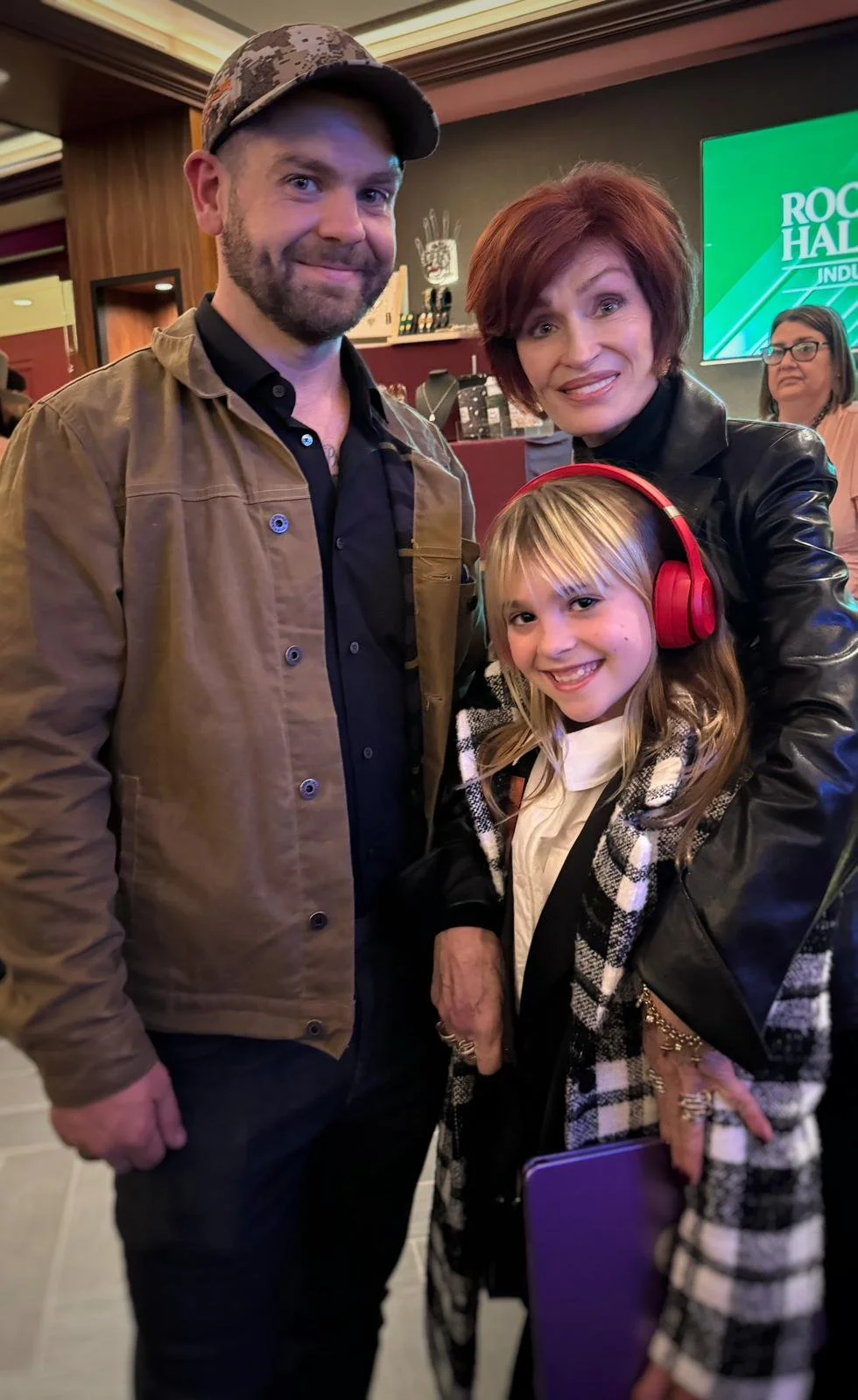 Osbourne family posing together indoors, with a man, a woman, and a girl wearing red headphones, all smiling. The background shows a bar area and a large green sign that says 'Rock Hall Induction.'