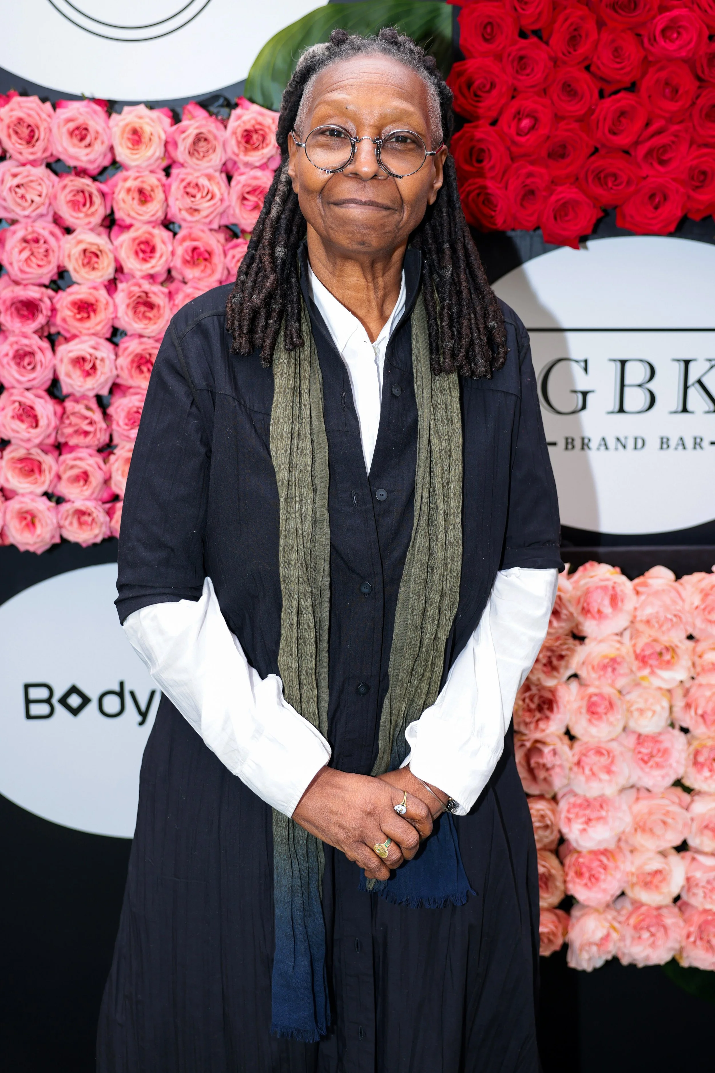 A woman with glasses and dreadlocks standing in front of a floral backdrop of pink and red roses.