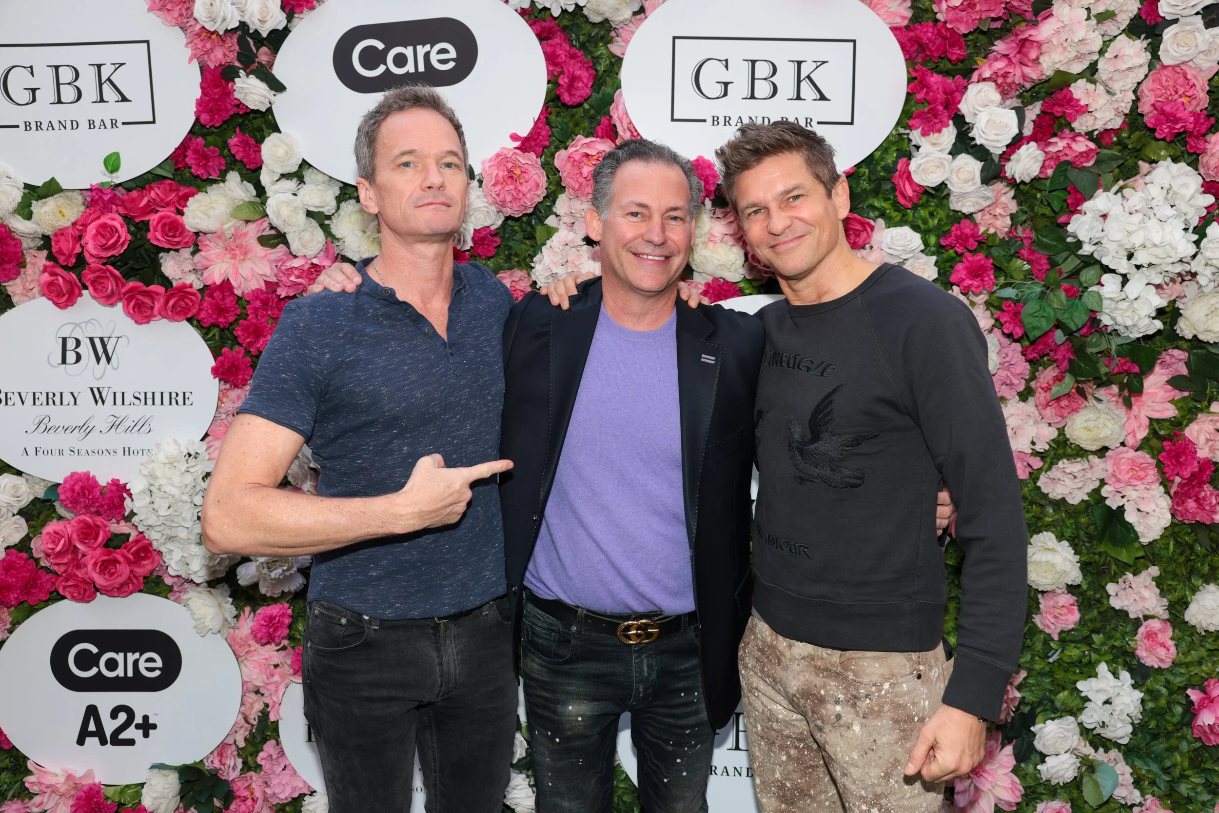 Three men standing together in front of a floral backdrop with logos, smiling and posing for the camera.