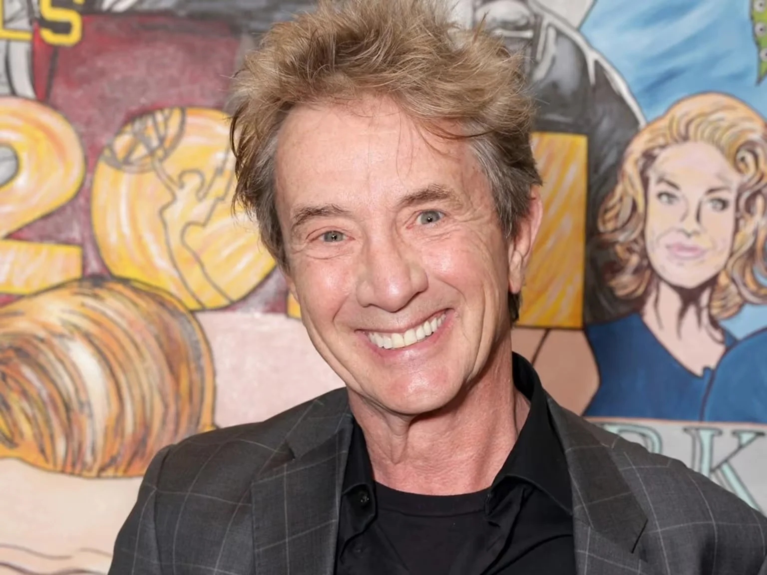 Smiling man with light brown hair wearing a dark checkered suit, standing in front of colorful artwork including a woman with blonde hair and abstract shapes.