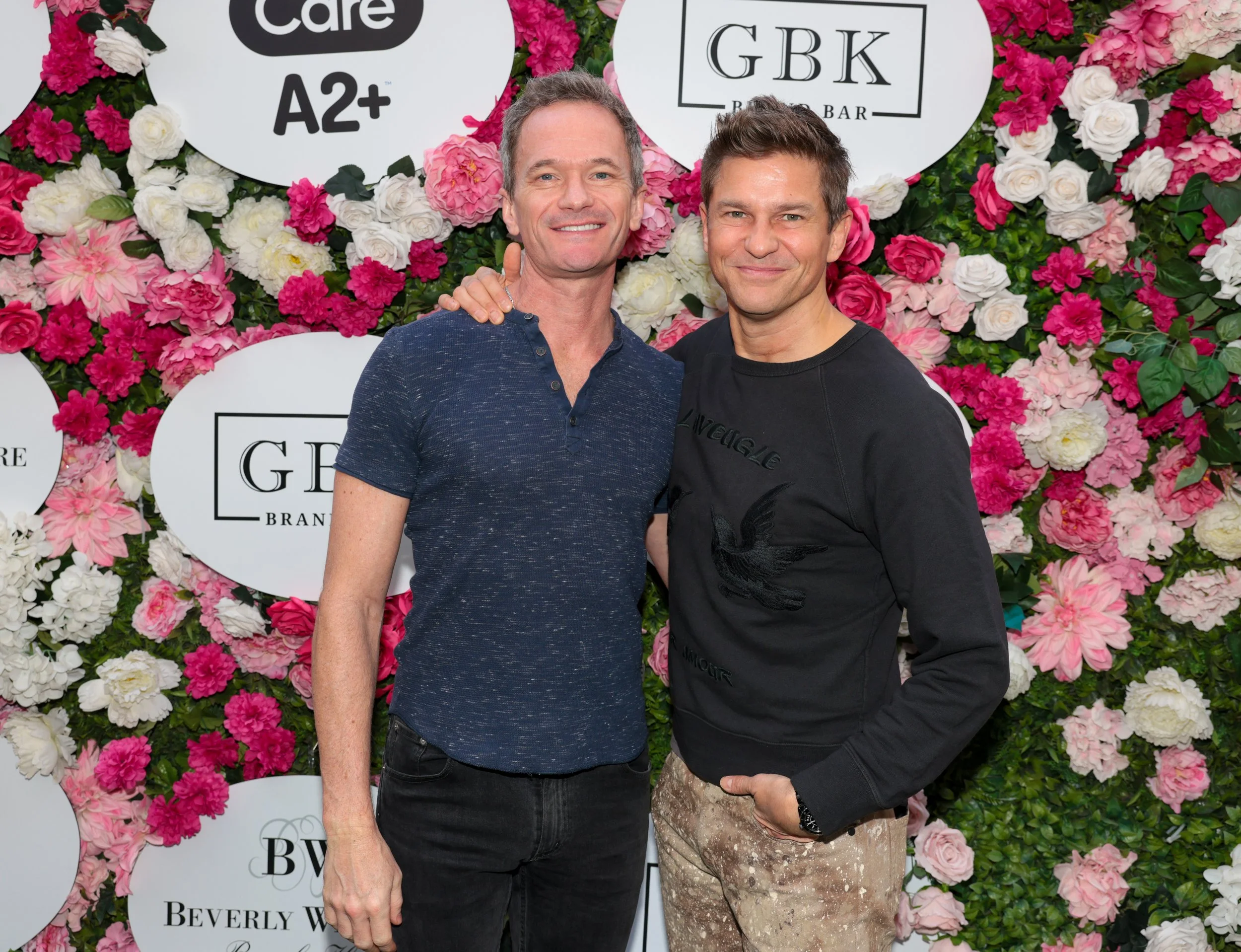 Two men smiling and standing close together in front of a floral backdrop with signed logos, one wearing a navy blue t-shirt and the other in a black sweatshirt and beige patterned pants.