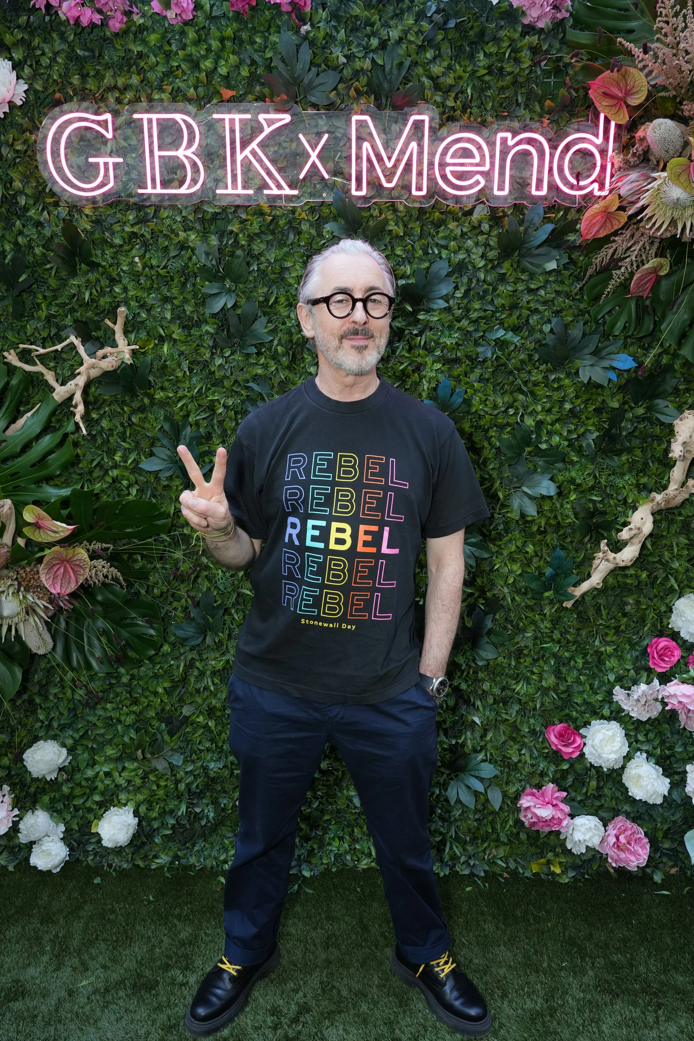 A man with gray hair and glasses making a peace sign stands in front of a lush green floral backdrop with pink and white flowers. A neon sign above him reads 'GBK x Mendl' in pink. The man is wearing a black T-shirt with colorful text that reads 'REBEL' repeatedly.