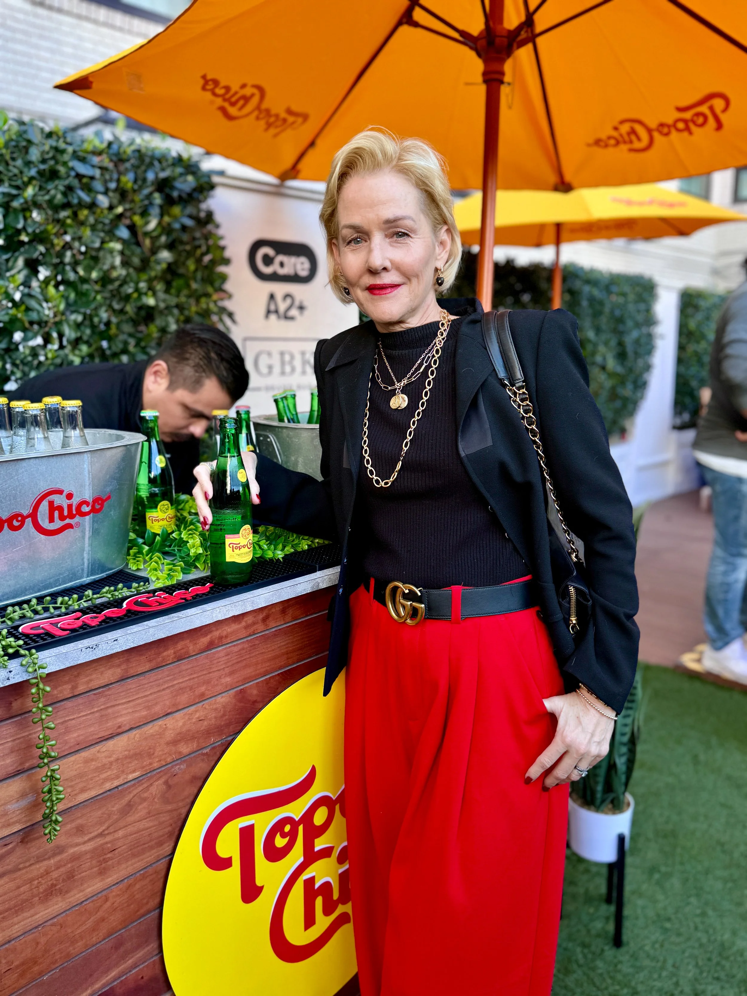 A woman with blonde hair wearing a black jacket, black top, and red pants, standing at a drink stand with a yellow umbrella that has the Topo Chico logo, holding a bottle of Topo Chico mineral water.