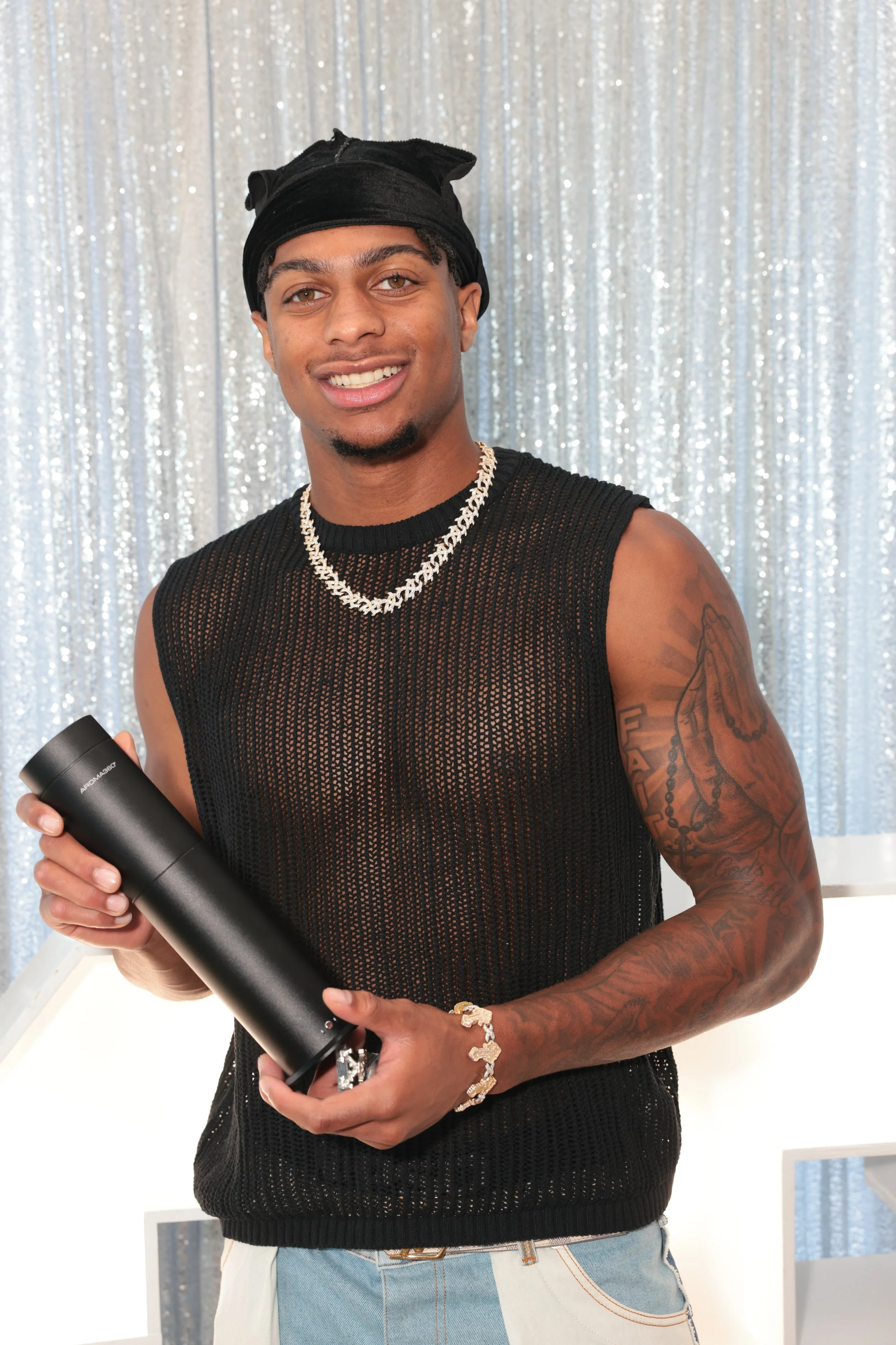 A young man with light brown skin, black dreadlocks, and a black headscarf, smiling and holding a black cylindrical object in a room with silver curtain background.