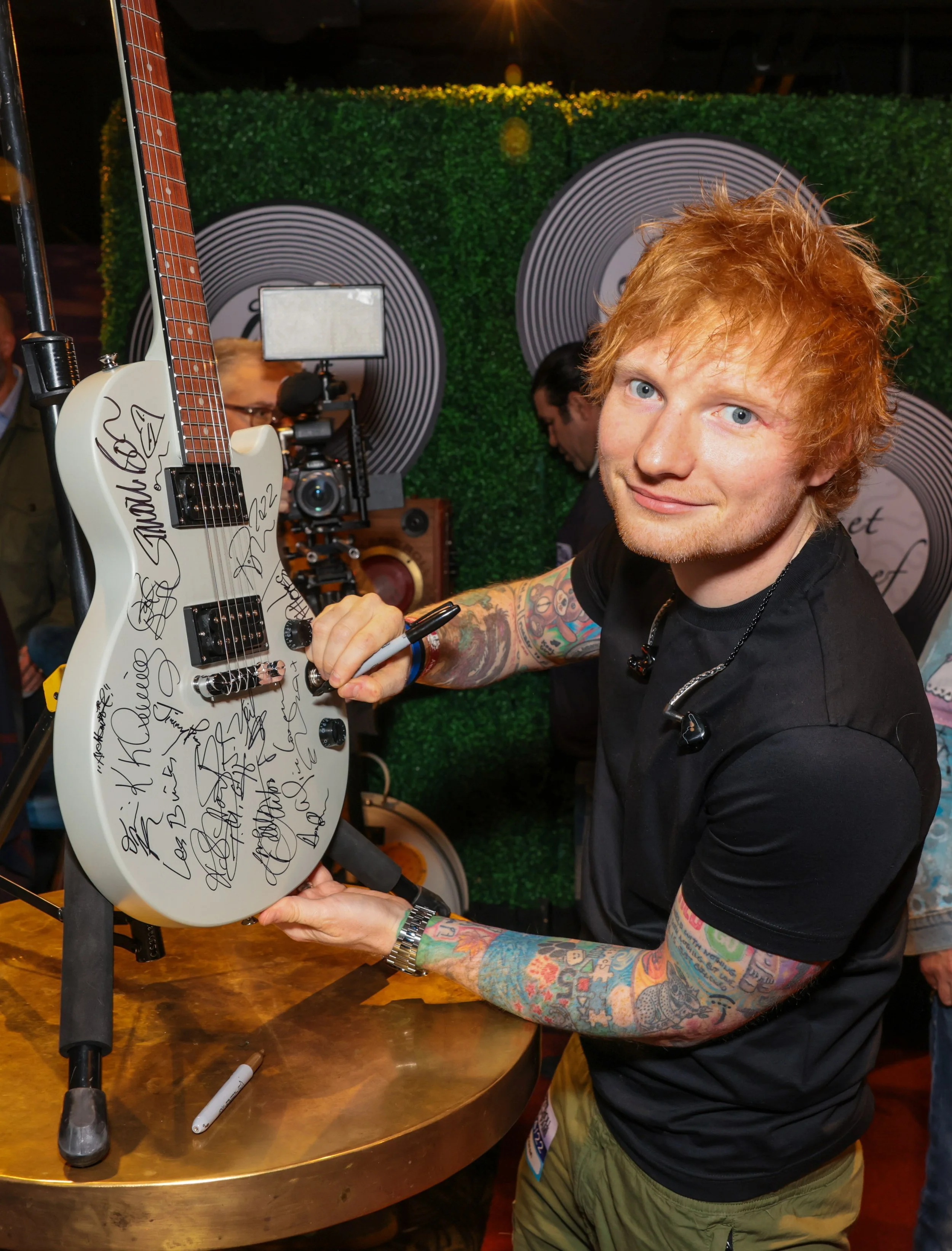 A man with orange hair and colorful tattoos on his arms is signing a white guitar covered in signatures. He is smiling at the camera at an event, with a green patterned wall in the background.