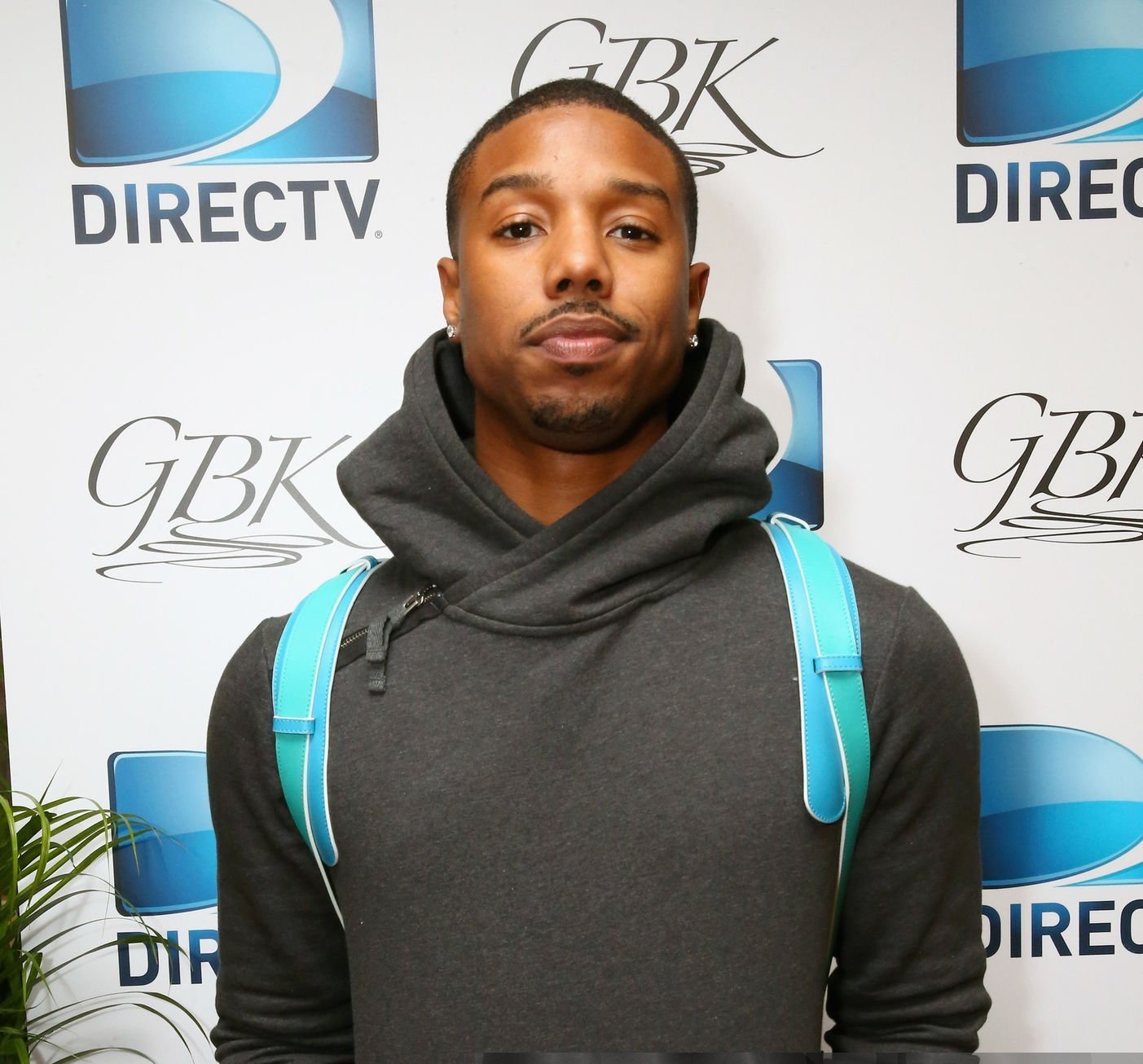 A young man with short hair, earrings, wearing a dark hoodie and carrying a light blue backpack, poses in front of a backdrop with 'DIRECTV' and 'GBK' logos.