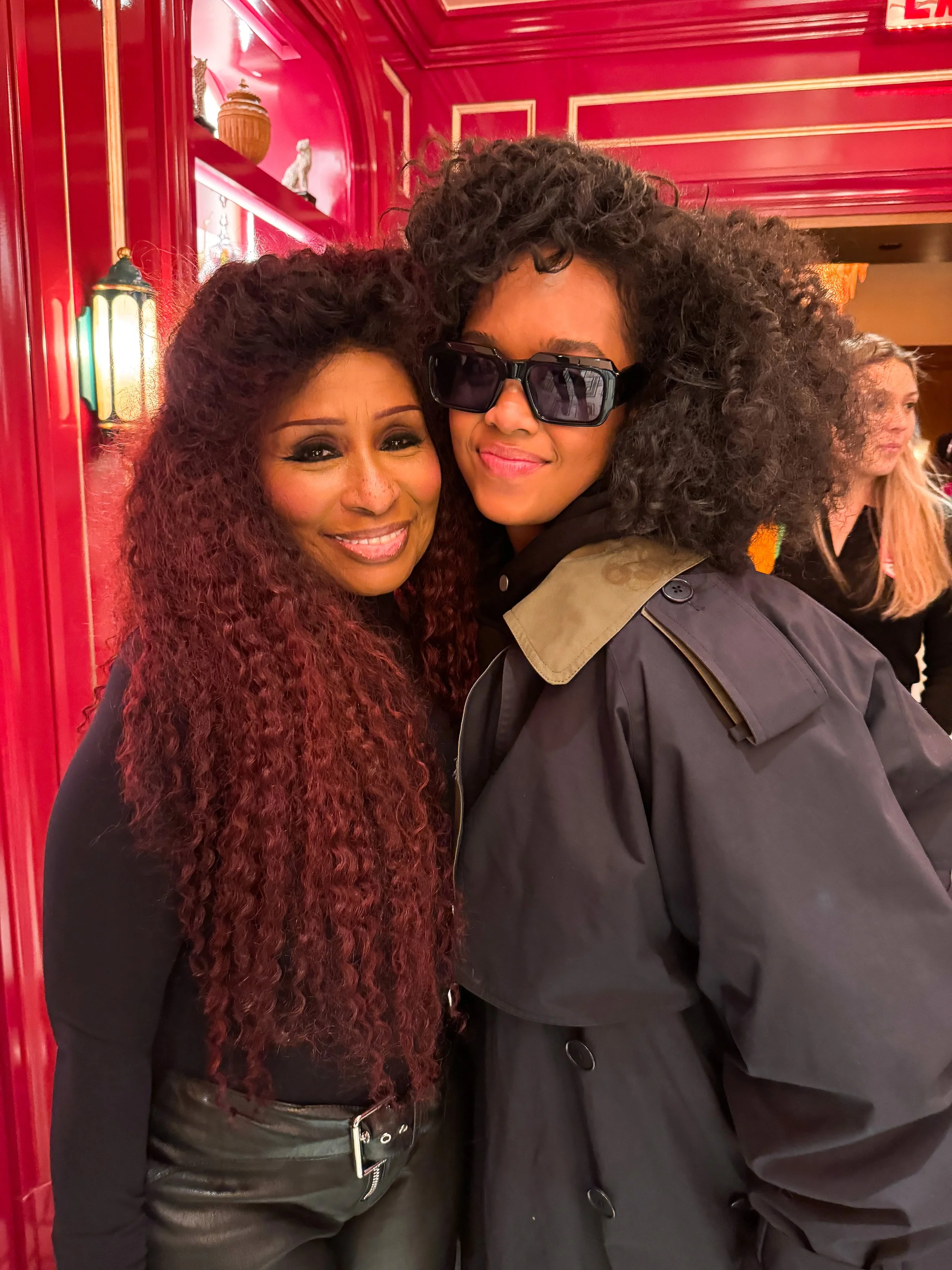 Two women with curly hair posing together inside a red, decorative room. One woman is smiling and has long, reddish-brown curly hair, wearing a black top. The other woman is wearing sunglasses, a black coat with a beige collar, and has voluminous curly hair.