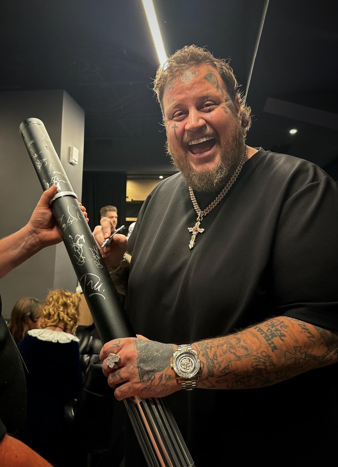 A man with tattoos, jewelry, and a beard is smiling as he holds a signed rolled poster or document. He is wearing a black shirt, a silver watch, and a chain with a cross pendant. The background shows people seated indoors, with dark walls and ceiling lights.