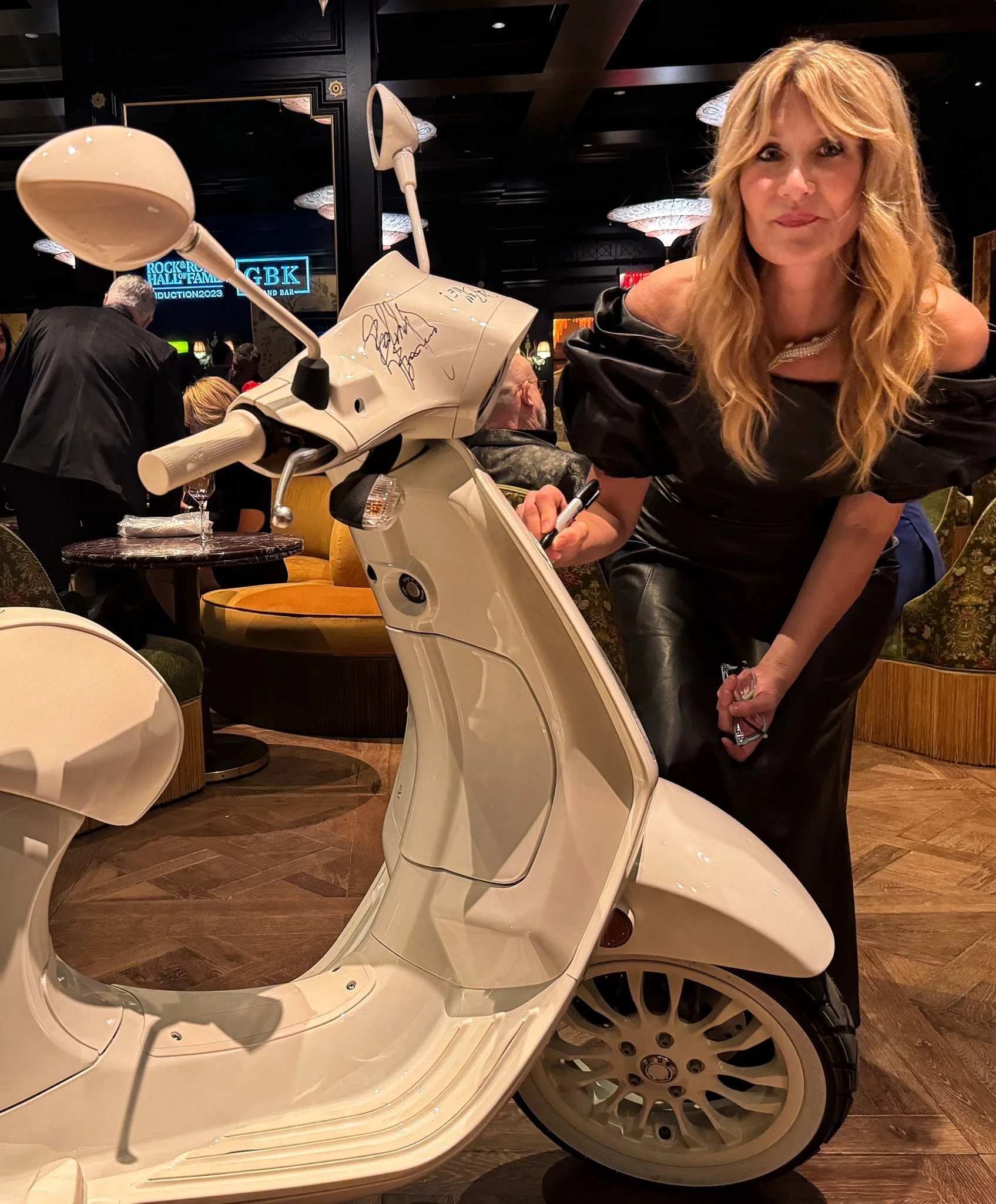 Woman with long wavy blonde hair leaning over a white scooter inside a dimly lit restaurant or lounge.