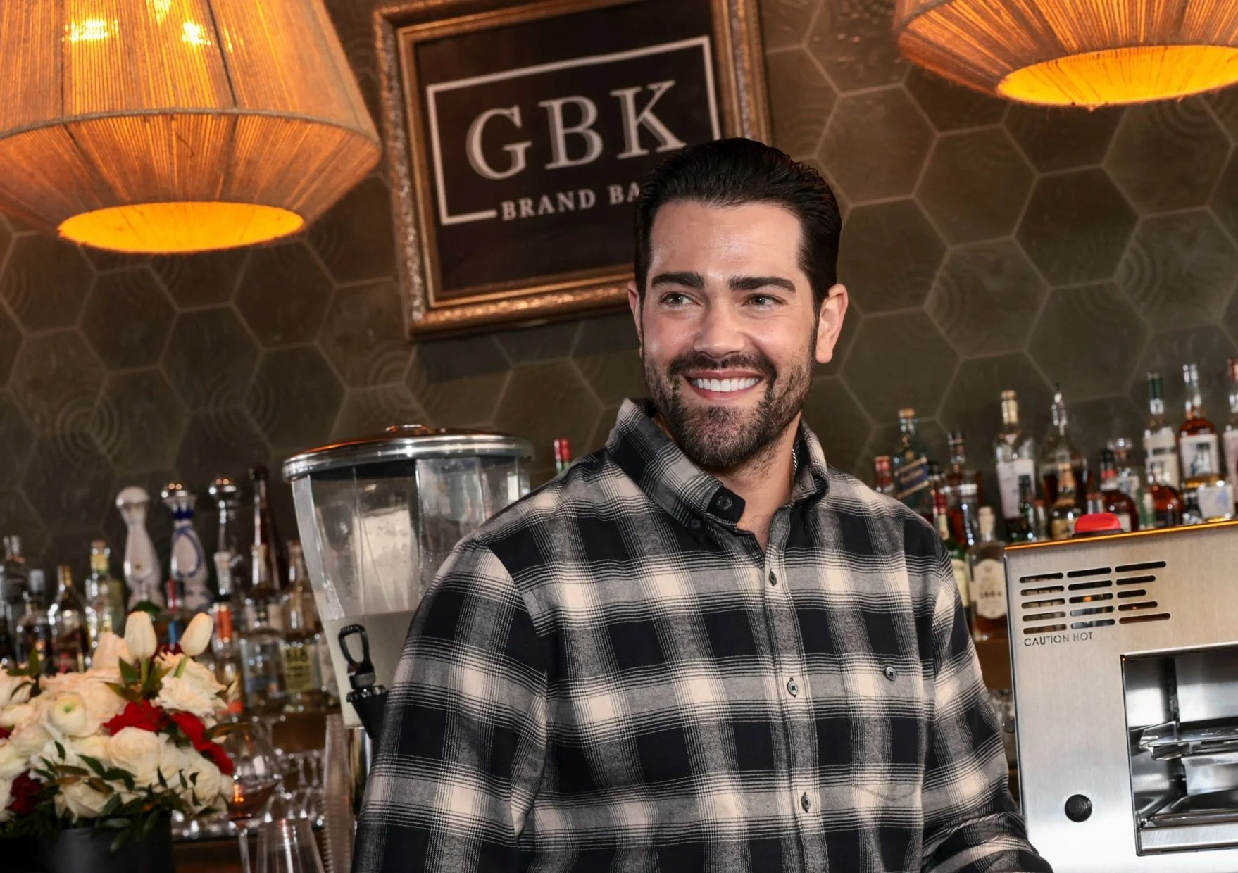 A smiling man with dark hair and a beard, wearing a black and gray plaid shirt, standing behind a bar with bottles and a blender in the background.
