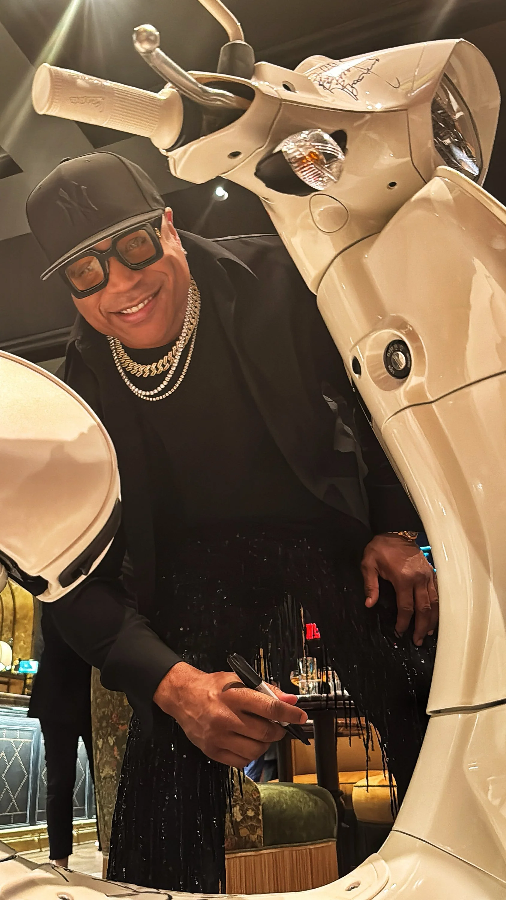 A man wearing a black cap, black glasses, a black shirt, and layered chains, smiling while signing an autograph on a white scooter's mirror with a marker inside a warmly lit, stylish room.
